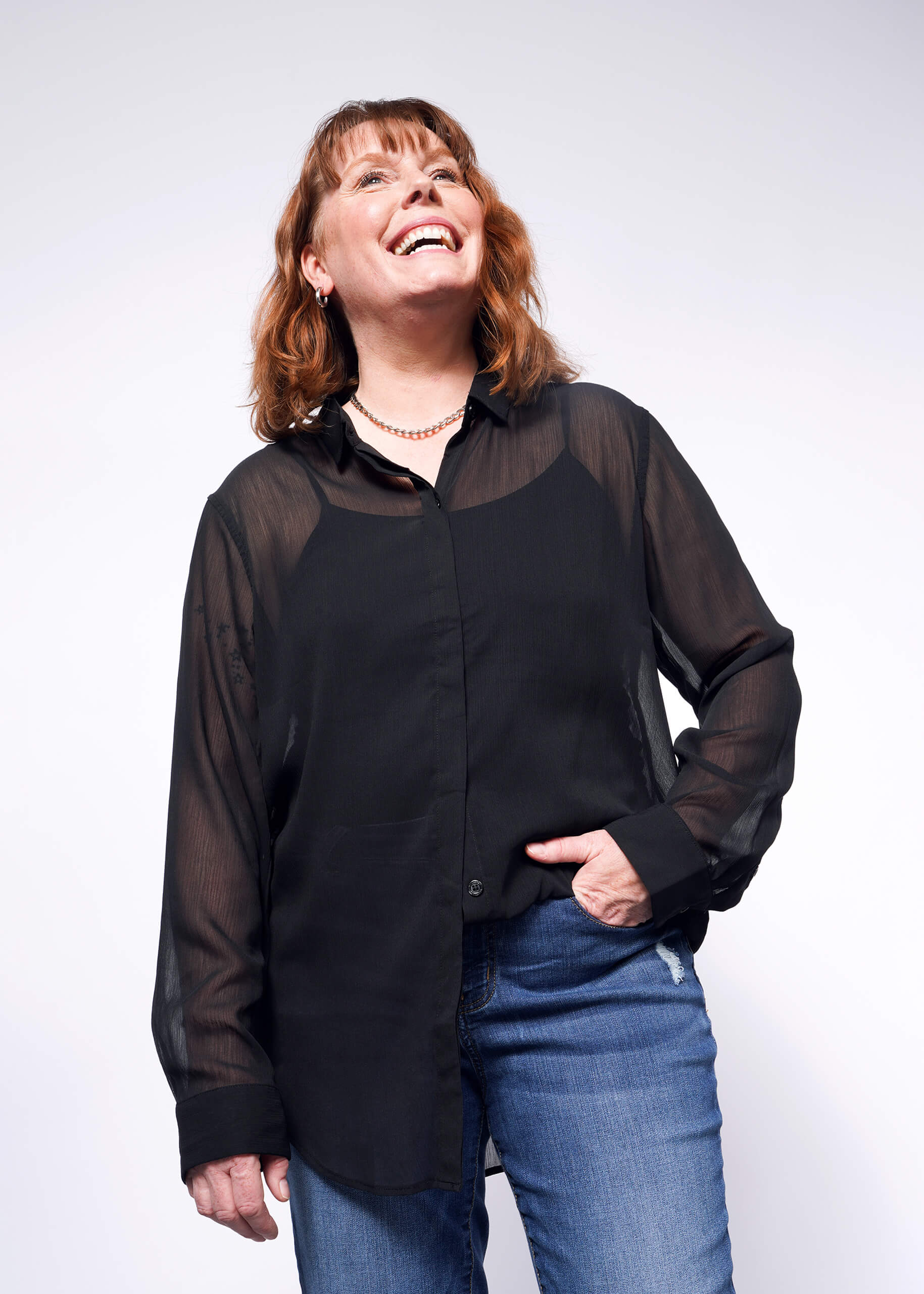 A woman with shoulder-length brown hair is joyfully looking up while wearing The Empower Sheer Long Sleeve Button Up, an oversized black top. She pairs it with blue jeans and her left hand in the pocket, all set against a plain white background.