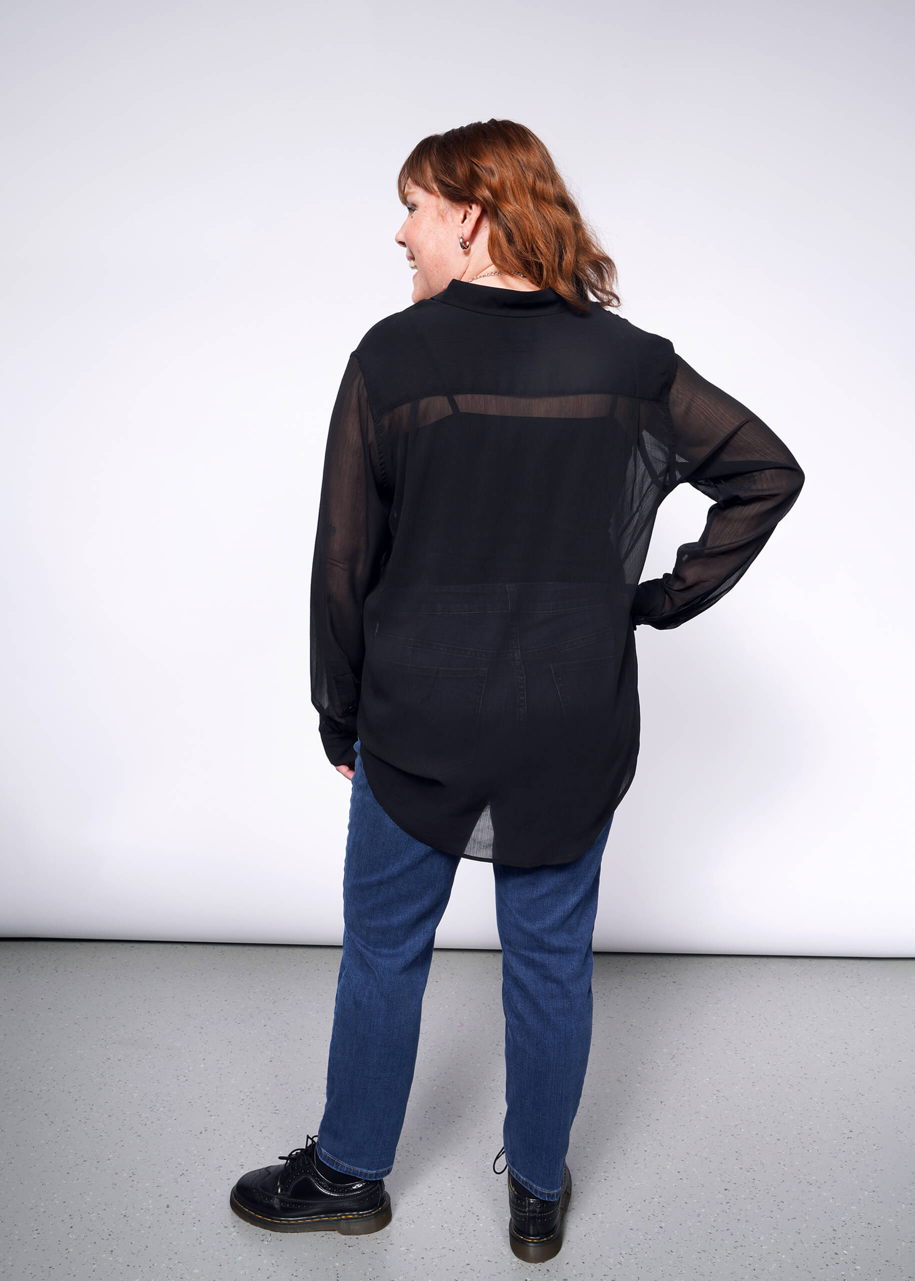 A person with shoulder-length brown hair stands facing a white wall, wearing The Empower Sheer Long Sleeve Button Up, blue jeans, and black shoes. Their left hand rests on their hip as they look slightly to the side.