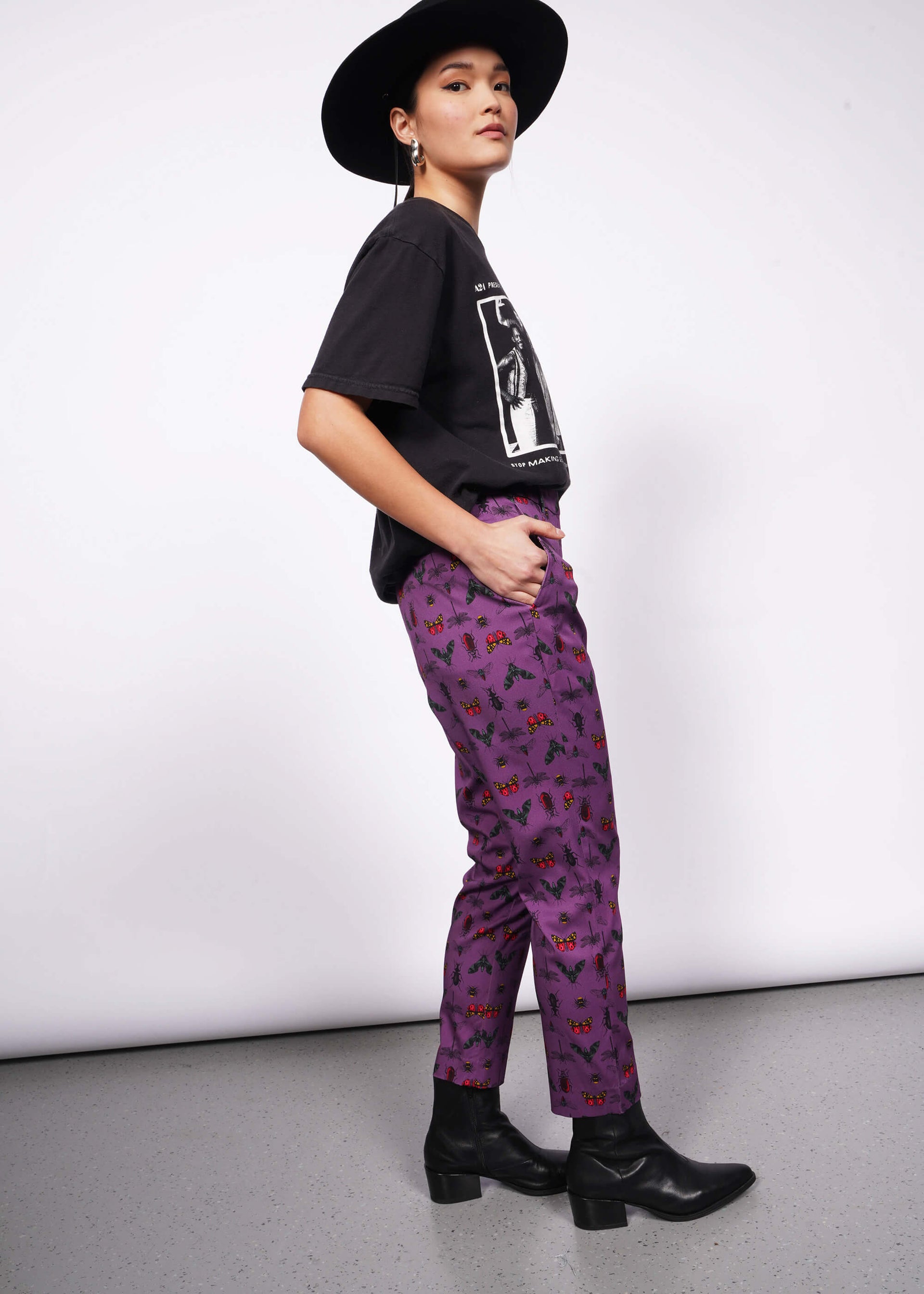 Side view of person wearing black wide brimmed hat, black band t-shirt and black heeled boots with hand in pocket of purple bug printed slim crop pants.