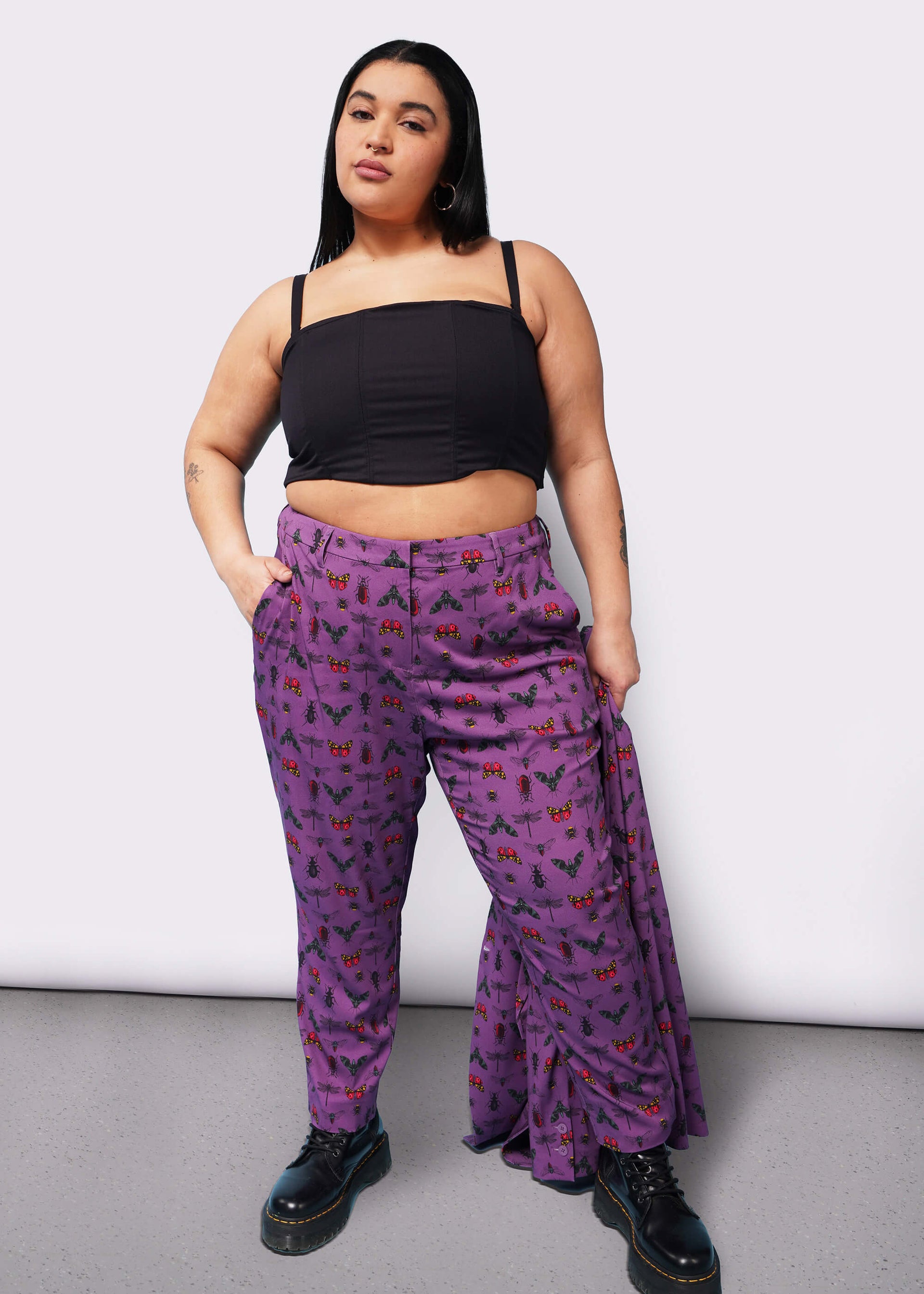 Person with long black hair looks at camera with hand in pocket wearing black corset, purple bug printed slim crop pants, and Doc Marten boots.