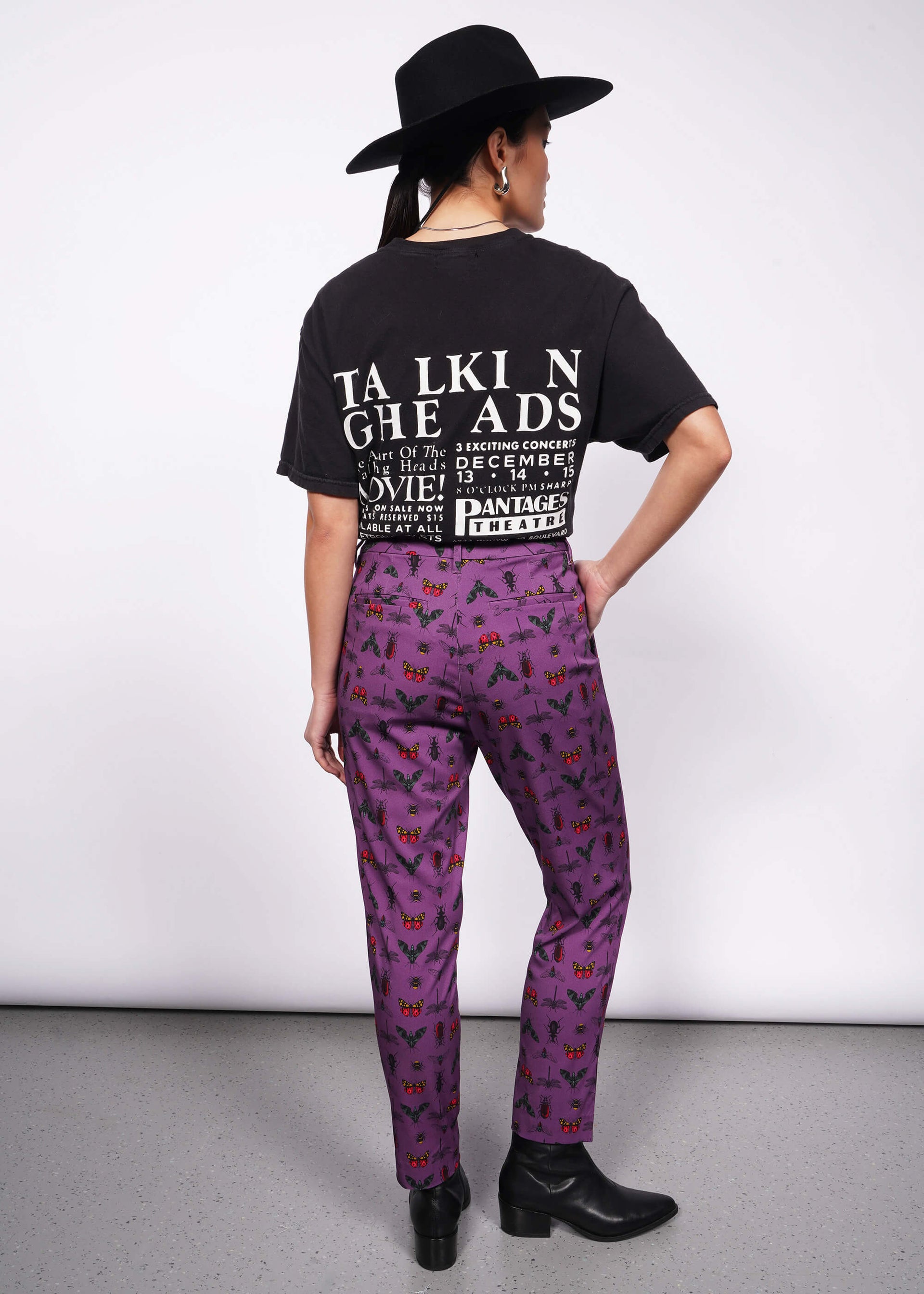 Back view of person wearing black wide brimmed hat, black band t-shirt and black heeled boots with hand in pocket of purple bug printed slim crop pants.