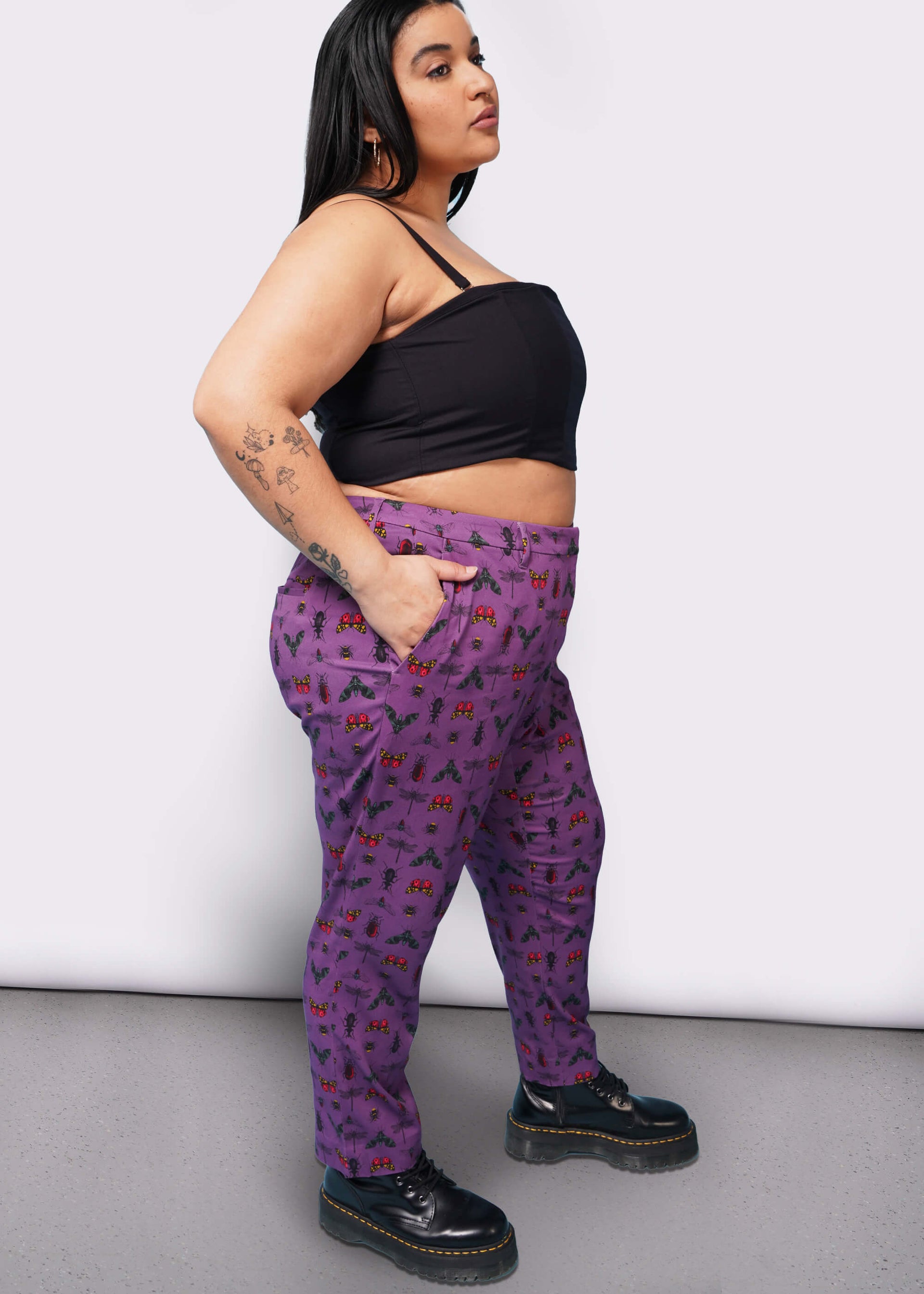 Side view of person with long black hair  with hand in pocket wearing black corset, purple bug printed slim crop pants, and Doc Marten boots.