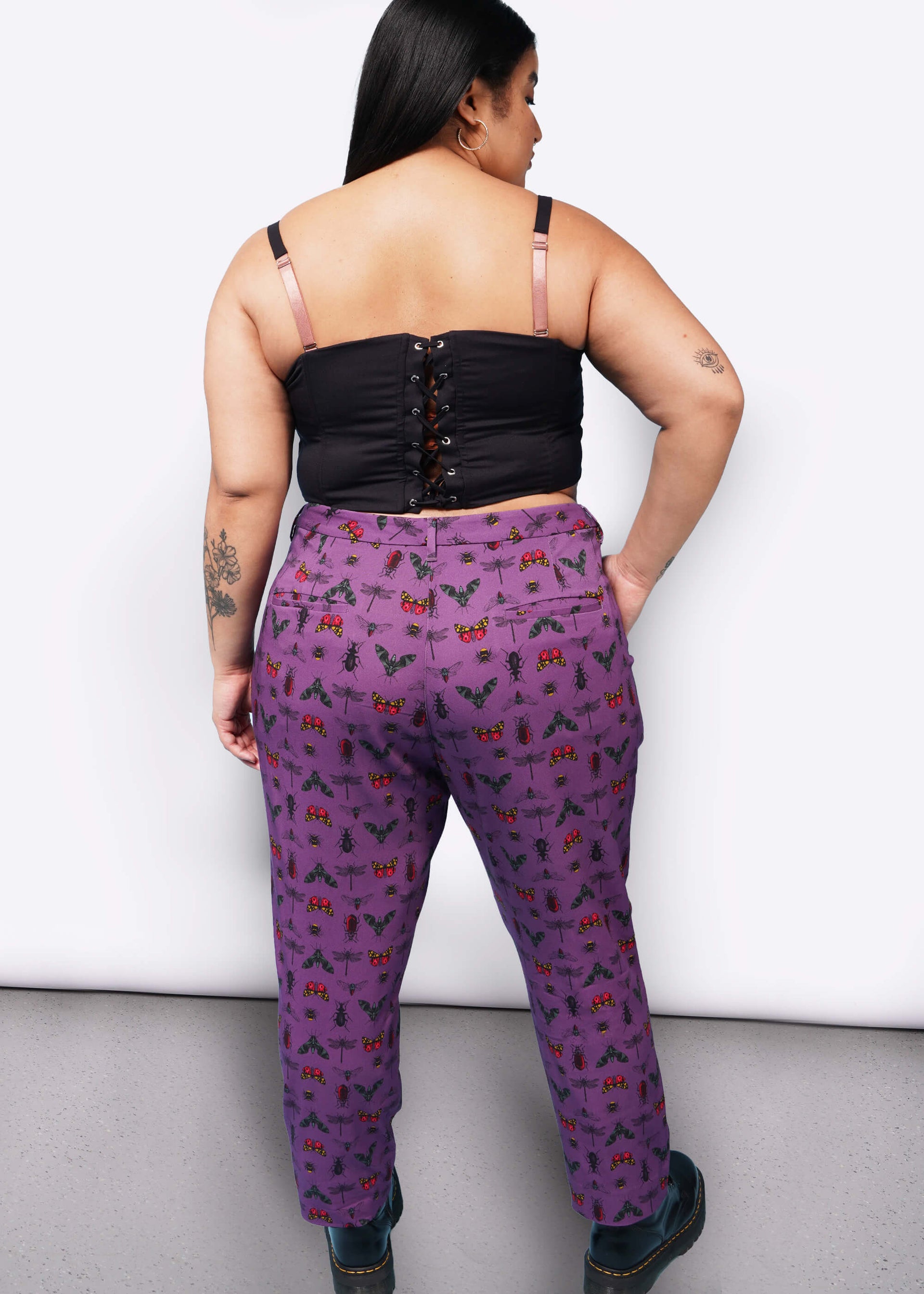 Back view of person with long black hair  with hand in pocket wearing black corset, purple bug printed slim crop pants, and Doc Marten boots.