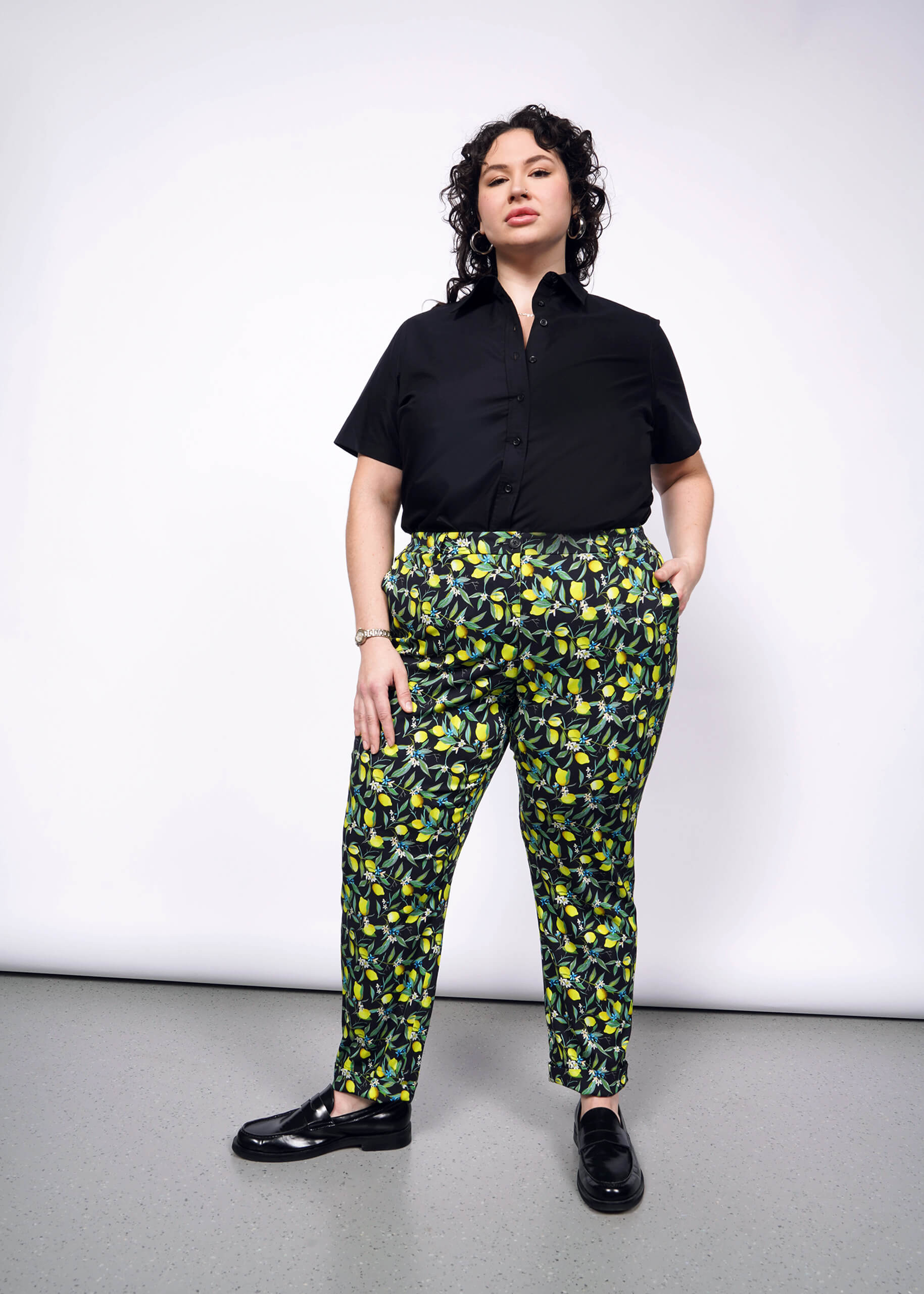 A person with curly black hair wearing a black shirt and vibrant yellow citrus printed pants, standing confidently with a smile.