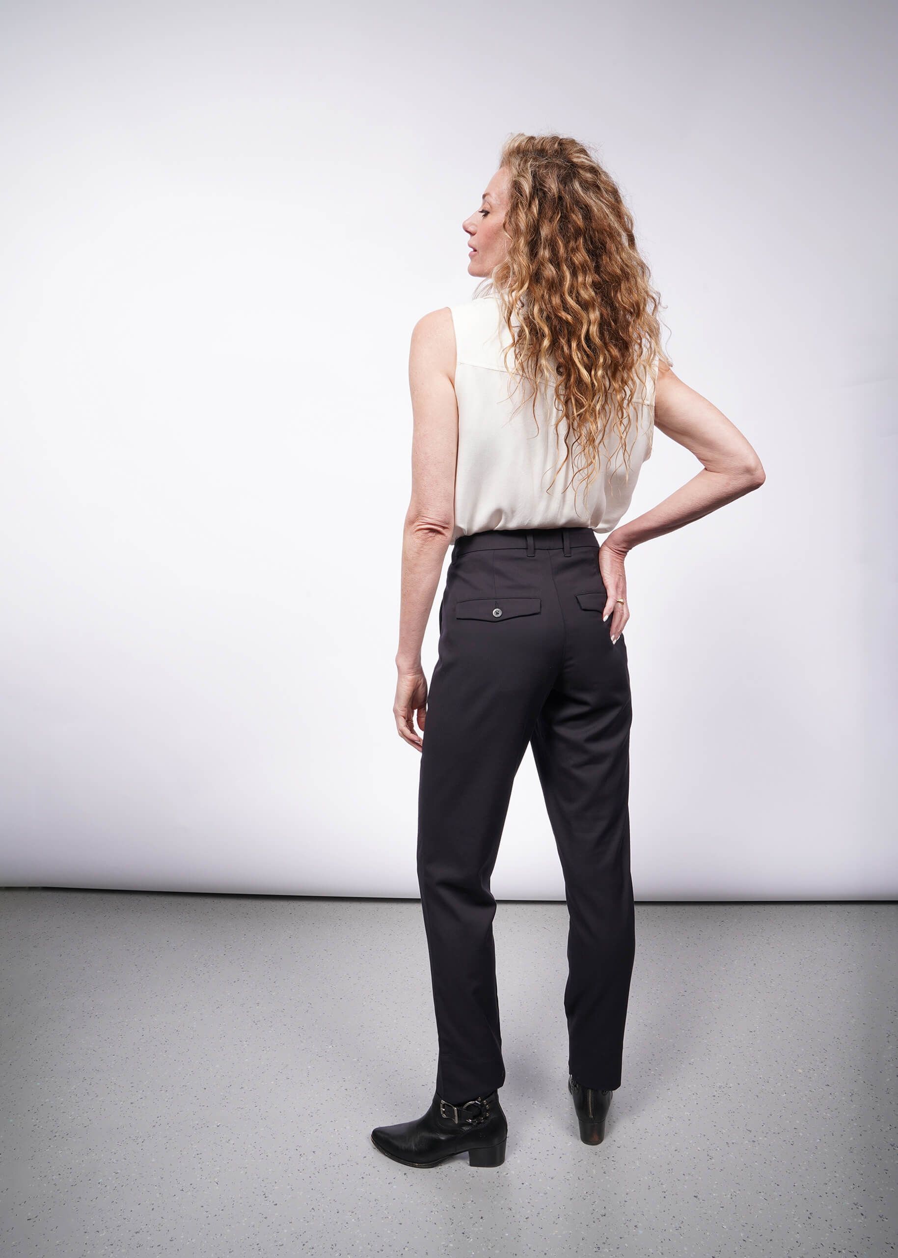 Facing sideways in a minimalist room, a person with long, curly hair wears a sleeveless white top paired with The Empower Trouser and matching black shoes. Their right hand rests on their hip against the backdrop of a plain, light gray wall.