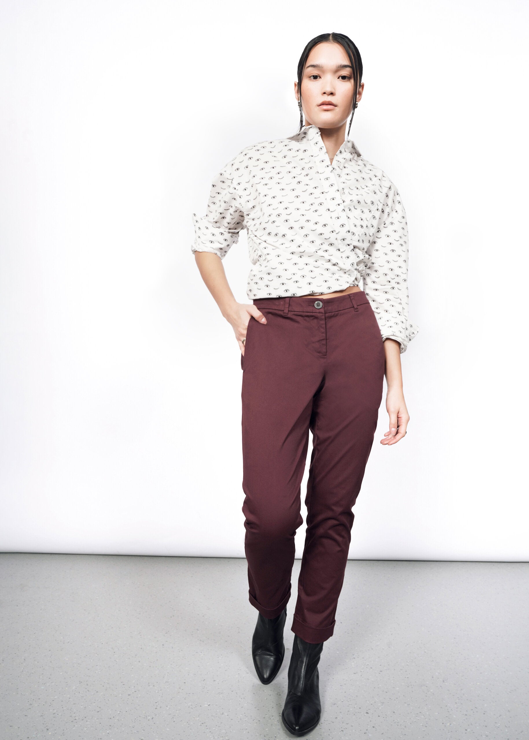 A person stands against a white backdrop wearing a patterned white shirt with convertible cuffs, The Essential Trouser in burgundy tapered-leg style, and black ankle boots. One hand is casually in a pocket, their expression neutral.