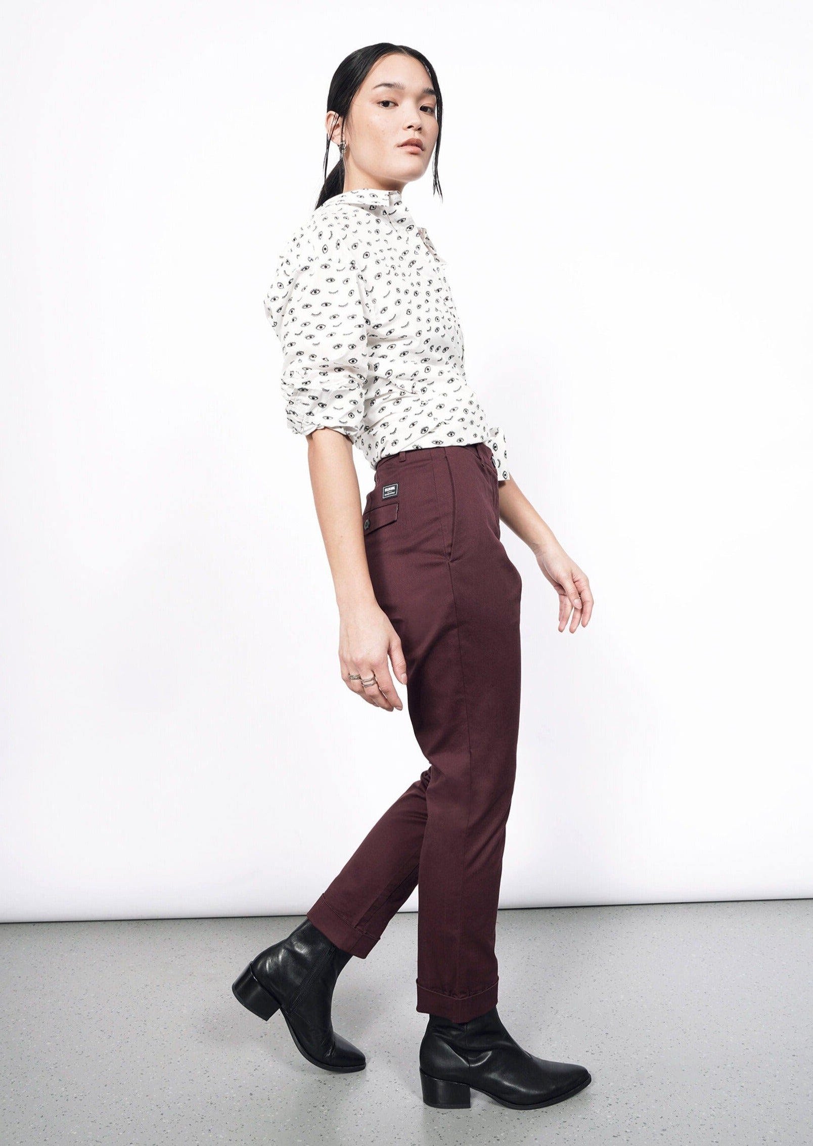 A person wearing a white patterned blouse with The Essential Trouser and black ankle boots poses sideways against a plain white background, gazing forward.