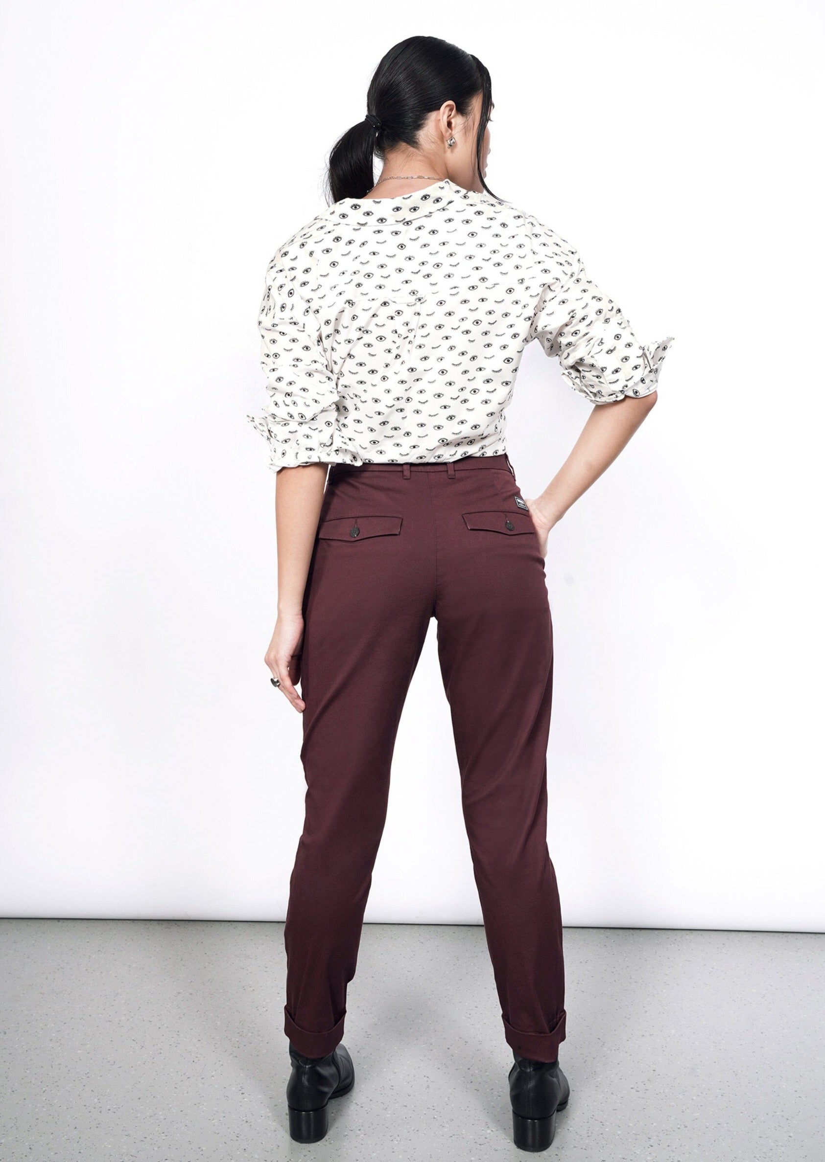 A woman stands facing away in The Essential Trouser, maroon with a tapered leg, paired with a white blouse featuring a black pattern. Her hair is tied back, and shes set against a plain white background on a speckled gray floor.