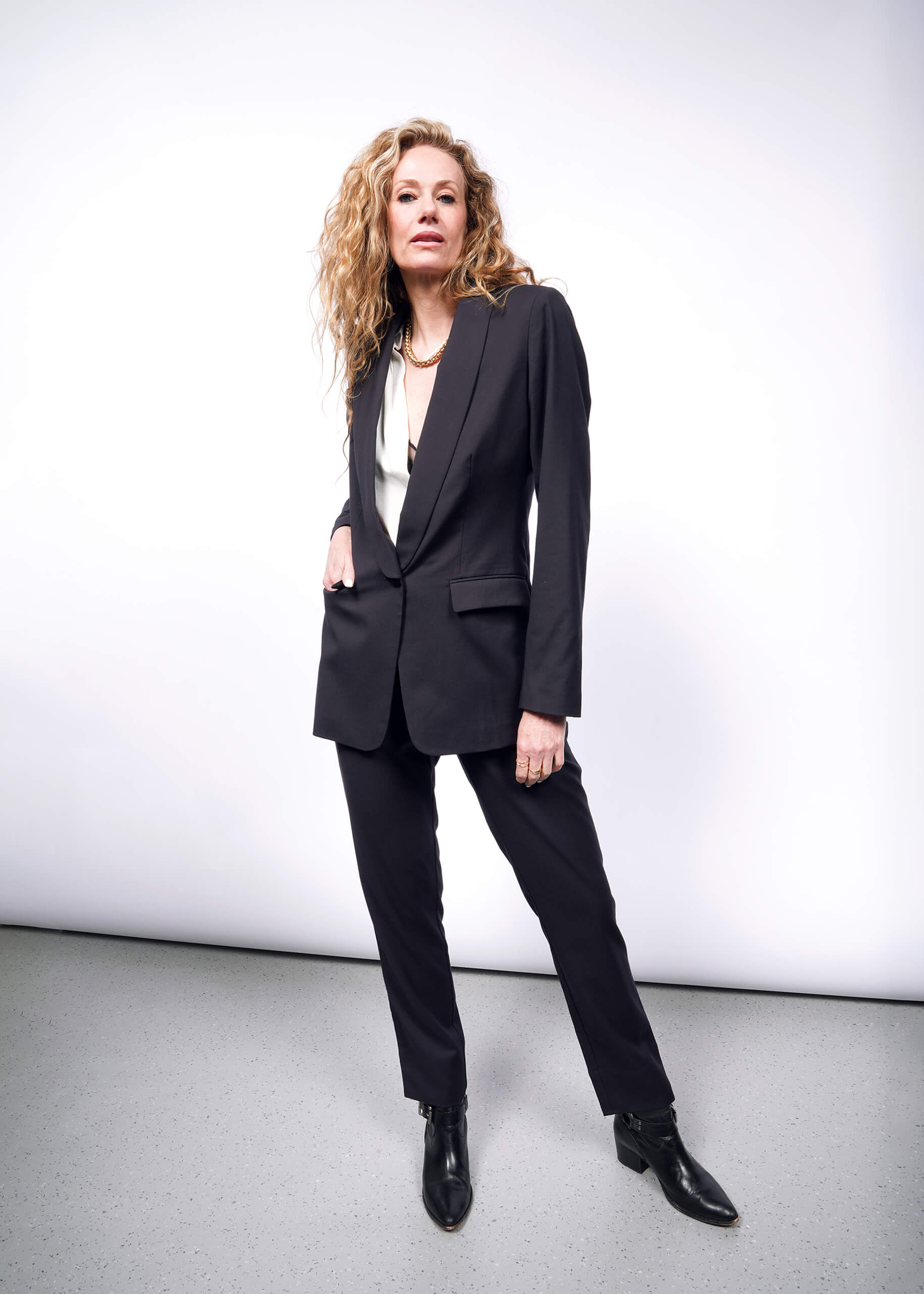 A person with long curly hair confidently stands against a plain white background in The Empower Tux Blazer, a black suit, and boots. Their left hand rests in the deep pocket of their pants, showcasing a relaxed yet assertive pose that complements the blazers timeless tailored fit.