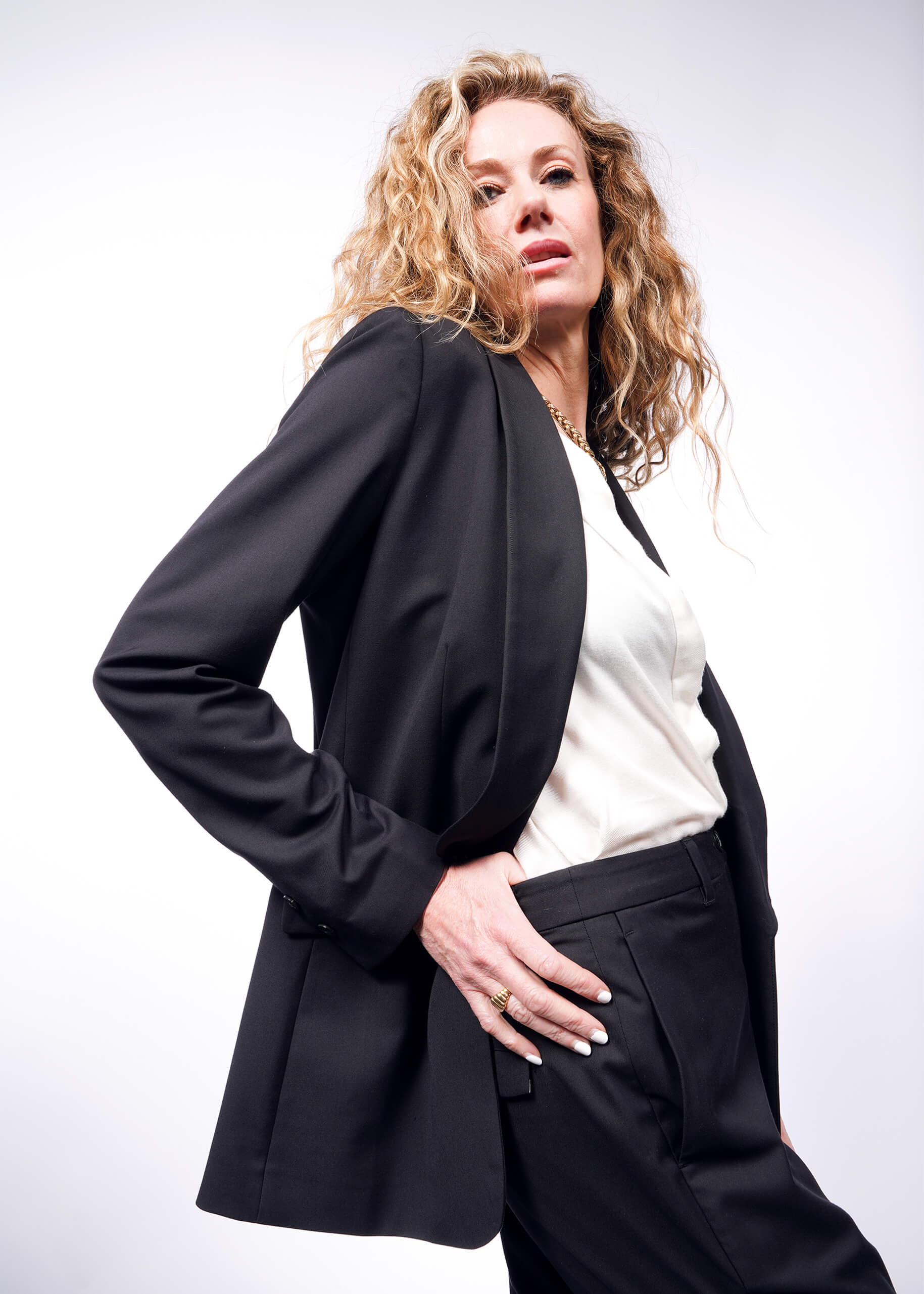 A woman with long curly hair exudes confidence in The Empower Tux Blazer. Posing against a white background, she wears a tailored black blazer and white top, hand on her hip while gazing slightly upwards.