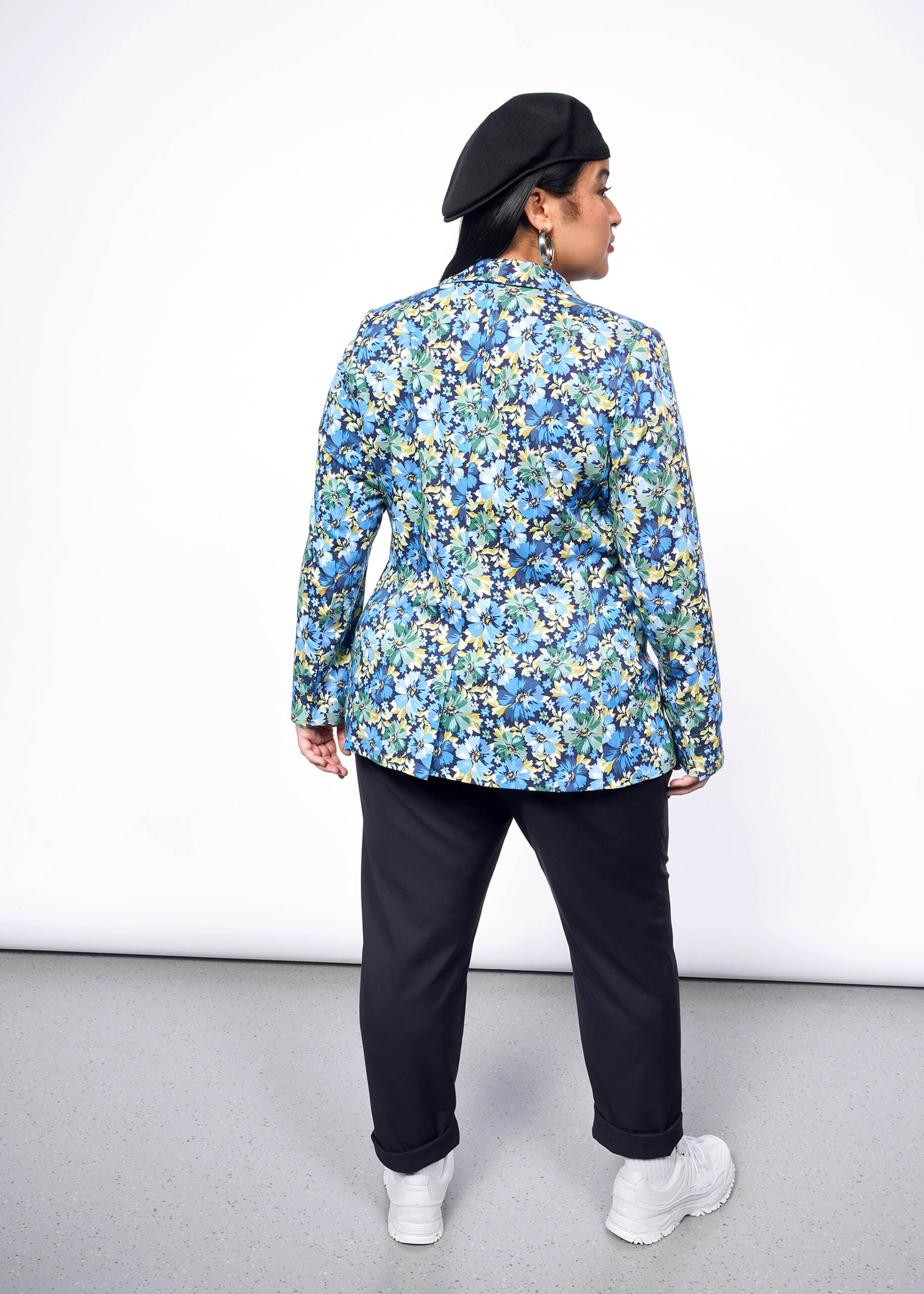 Back view of person with long black hair wearing Kangol hat, black pants, white sneakers, and floral patterned blazer.