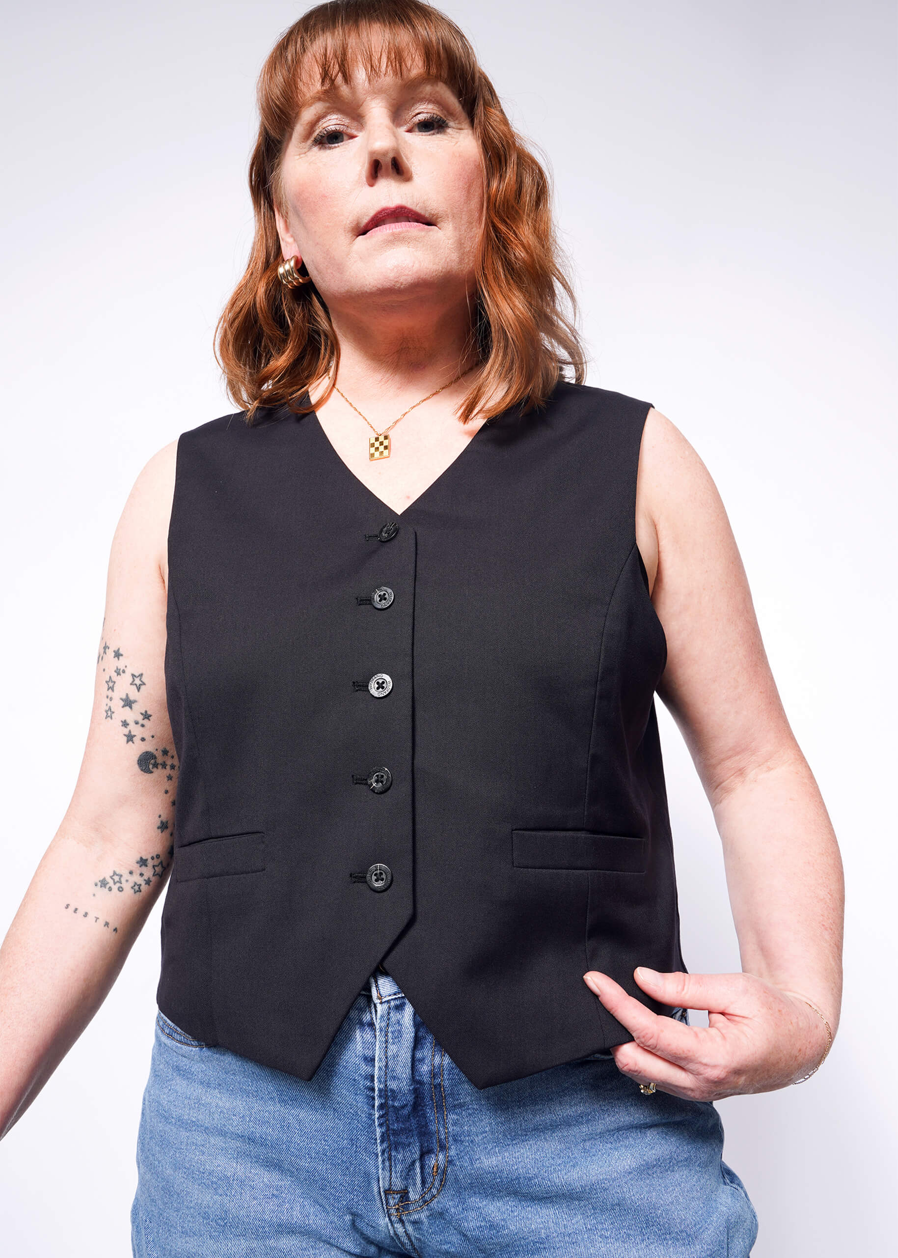 A person with long, reddish-brown hair stands confidently in The Empower Vest, blue jeans, and a necklace. Star tattoos adorn their right arm as they hold the vests hem against a plain backdrop.