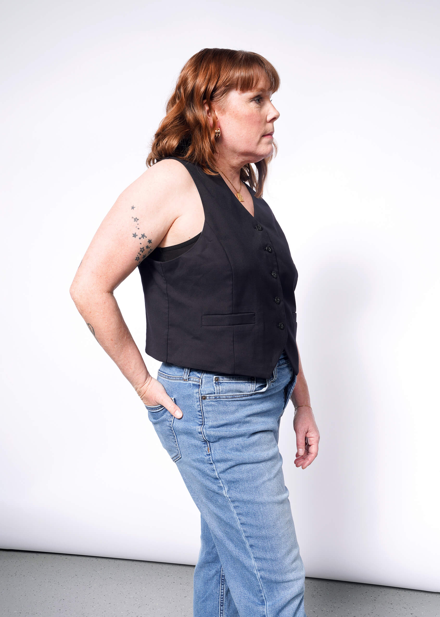 A person with shoulder-length hair stands against a plain background, wearing The Empower Vest over a white shirt and blue jeans. Their right hand is in their pocket, highlighting the tailored fit, while star tattoos adorn their left arm.