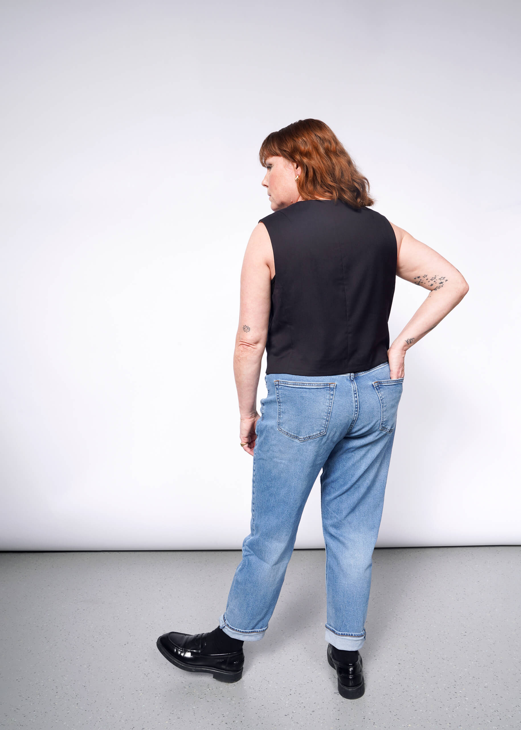 A person with shoulder-length hair wears The Empower Vest, a black sleeveless top with a tailored fit, over blue jeans and black shoes. They stand on a gray floor against a plain white background, facing away from the camera.