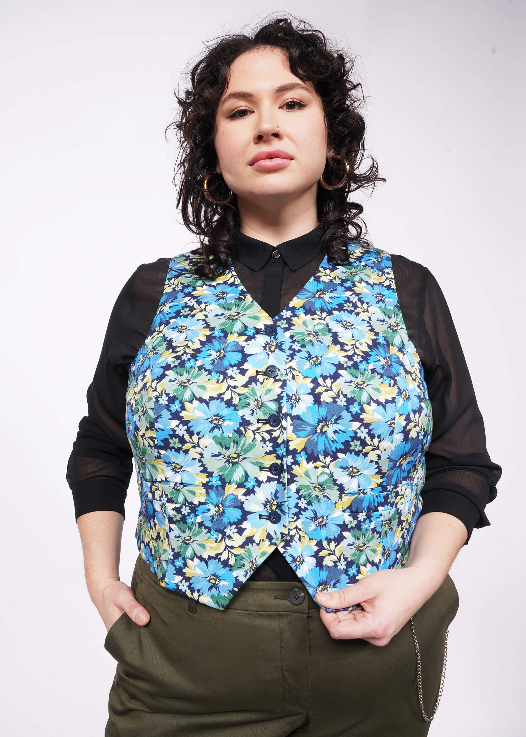 Person with short curly black hair looks at camera wearing black long sleeve button up, green pants, and floral patterned vest.