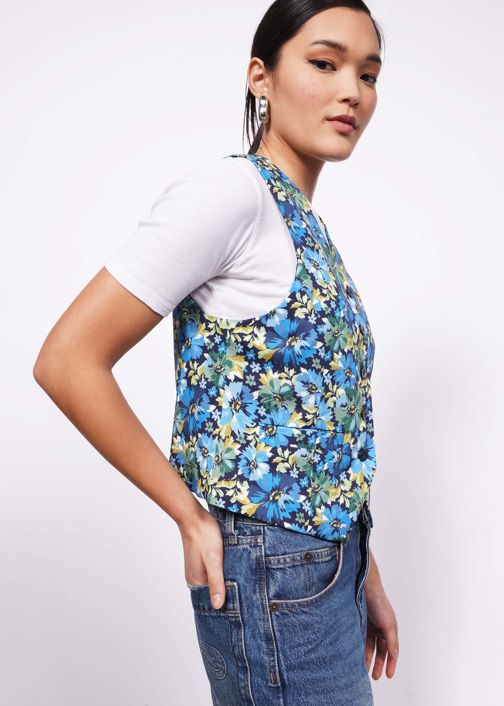 Side view of person with black hair tied back looking at camera with hand in back pocket wearing white tshirt, jeans, and a floral patterned vest.