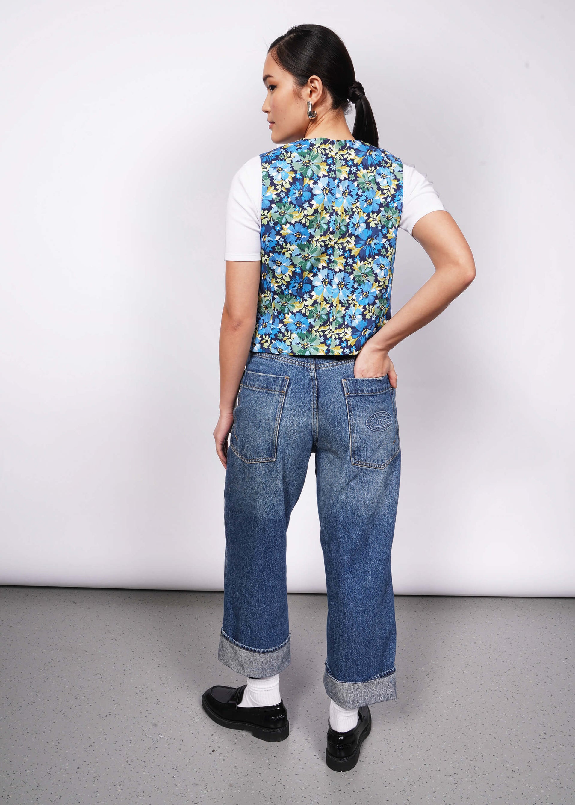 Back view of person with black hair tied back looking at camera with hand in back pocket wearing white tshirt, jeans, loafers, and a floral patterned vest.