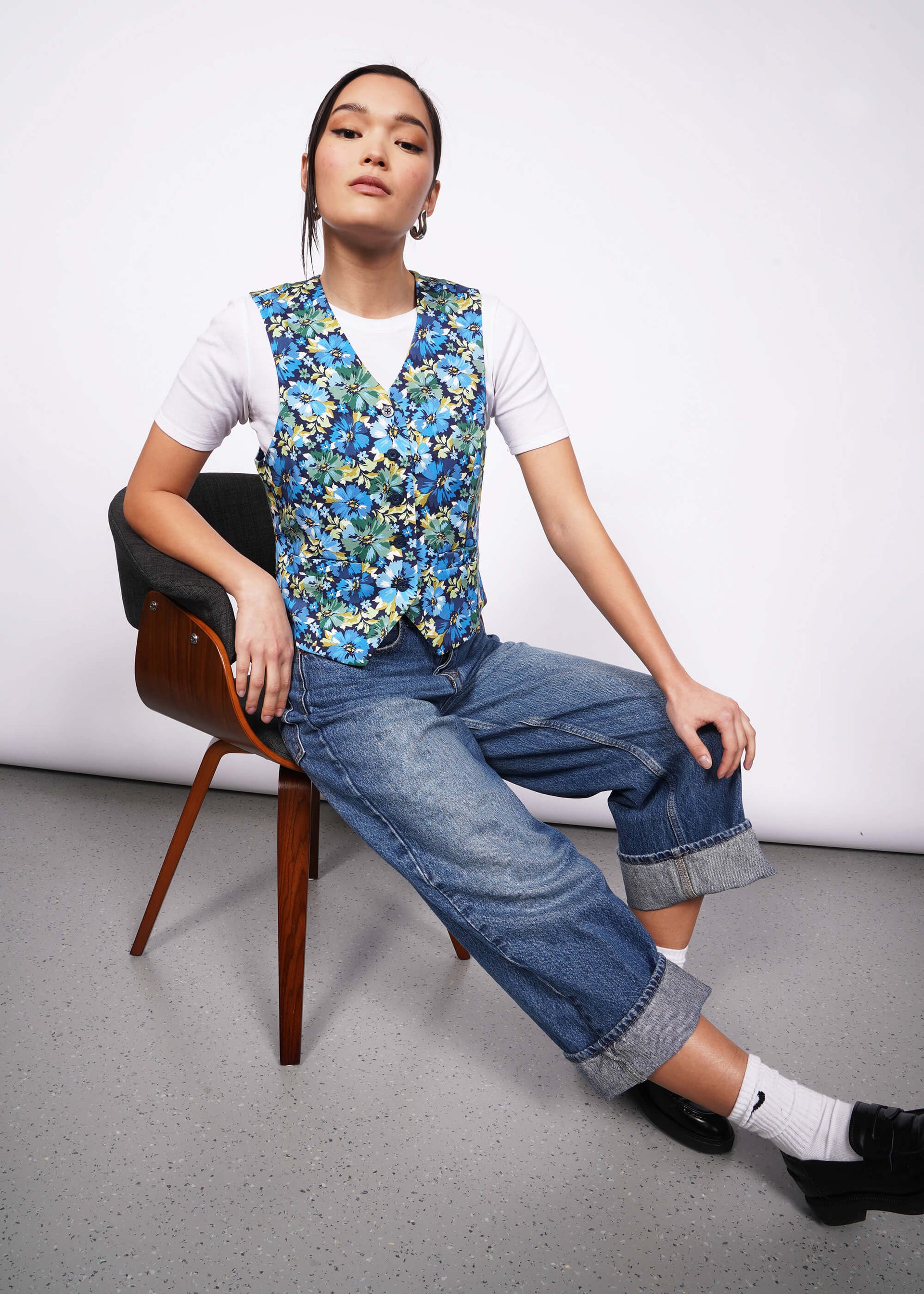 Person with black hair tied back sits on edge of chair with one leg extended looking at camera with hand on knee wearing white tshirt, jeans, loafers, and a floral patterned vest.