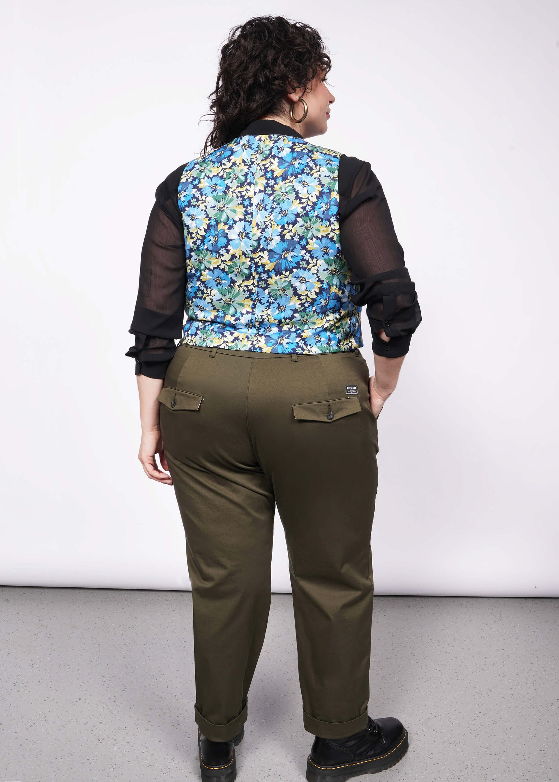 Back of person with short curly black hair wearing gold hoop earrings, a black button up shirt, green trousers, Doc Marten boots, and a floral printed vest.