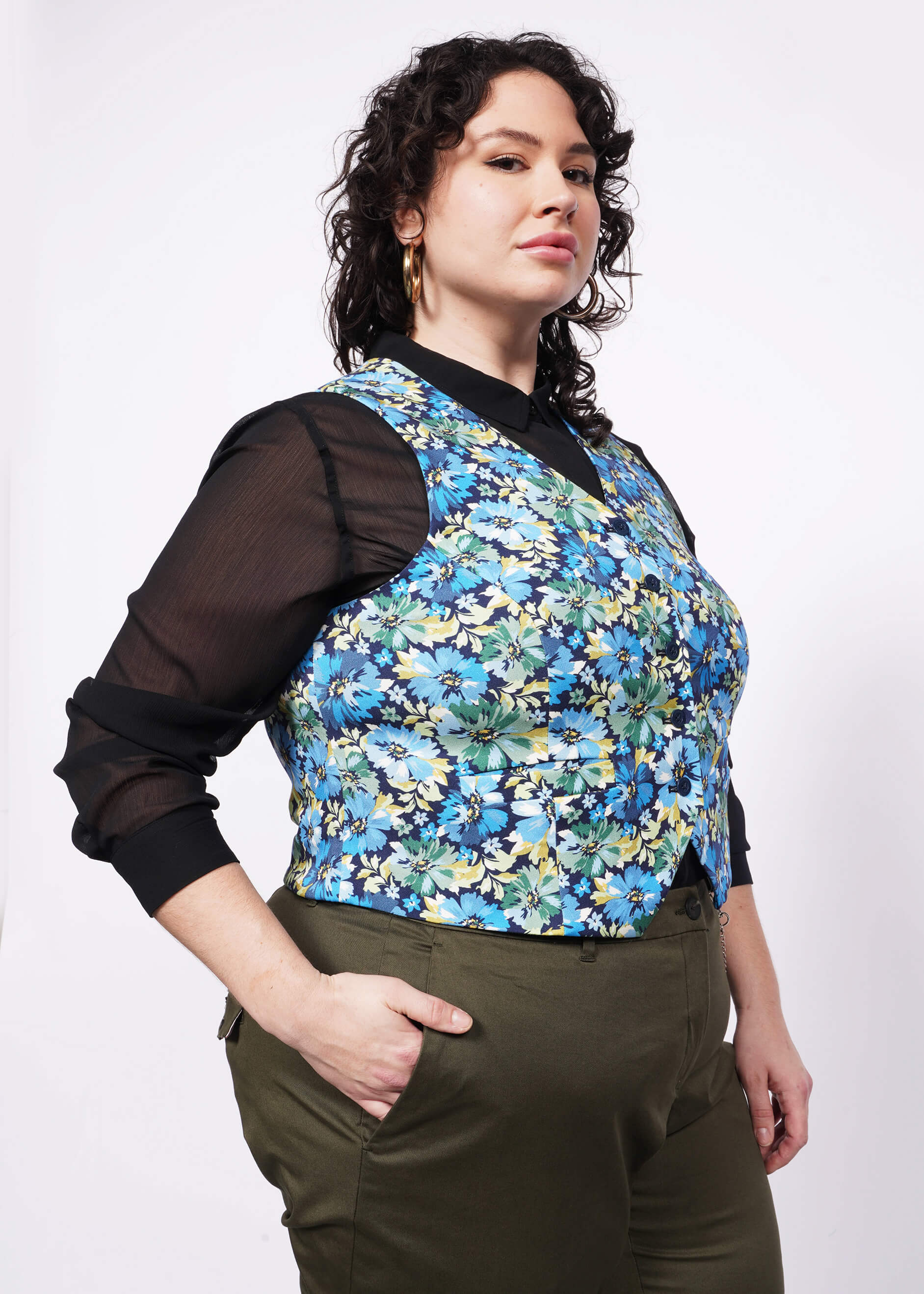 Side view of person with short curly black hair looking at camera with hand in pocket wearing gold hoop earrings, a black button up shirt, green trousers, and an unbuttoned floral printed vest.