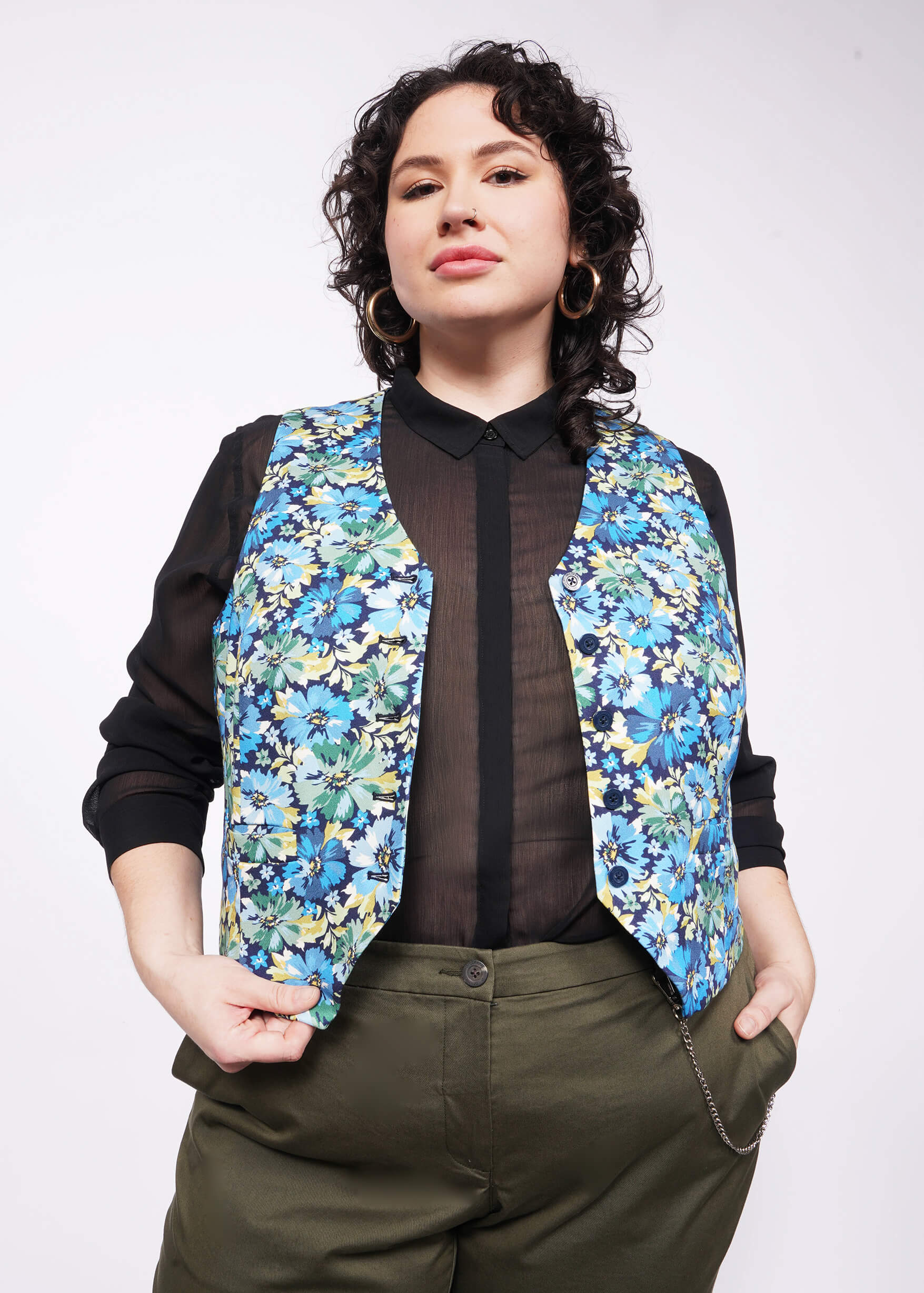 Person with short curly black hair looks at camera wearing gold hoop earrings, a black button up shirt, green trousers, and an unbuttoned floral printed vest.