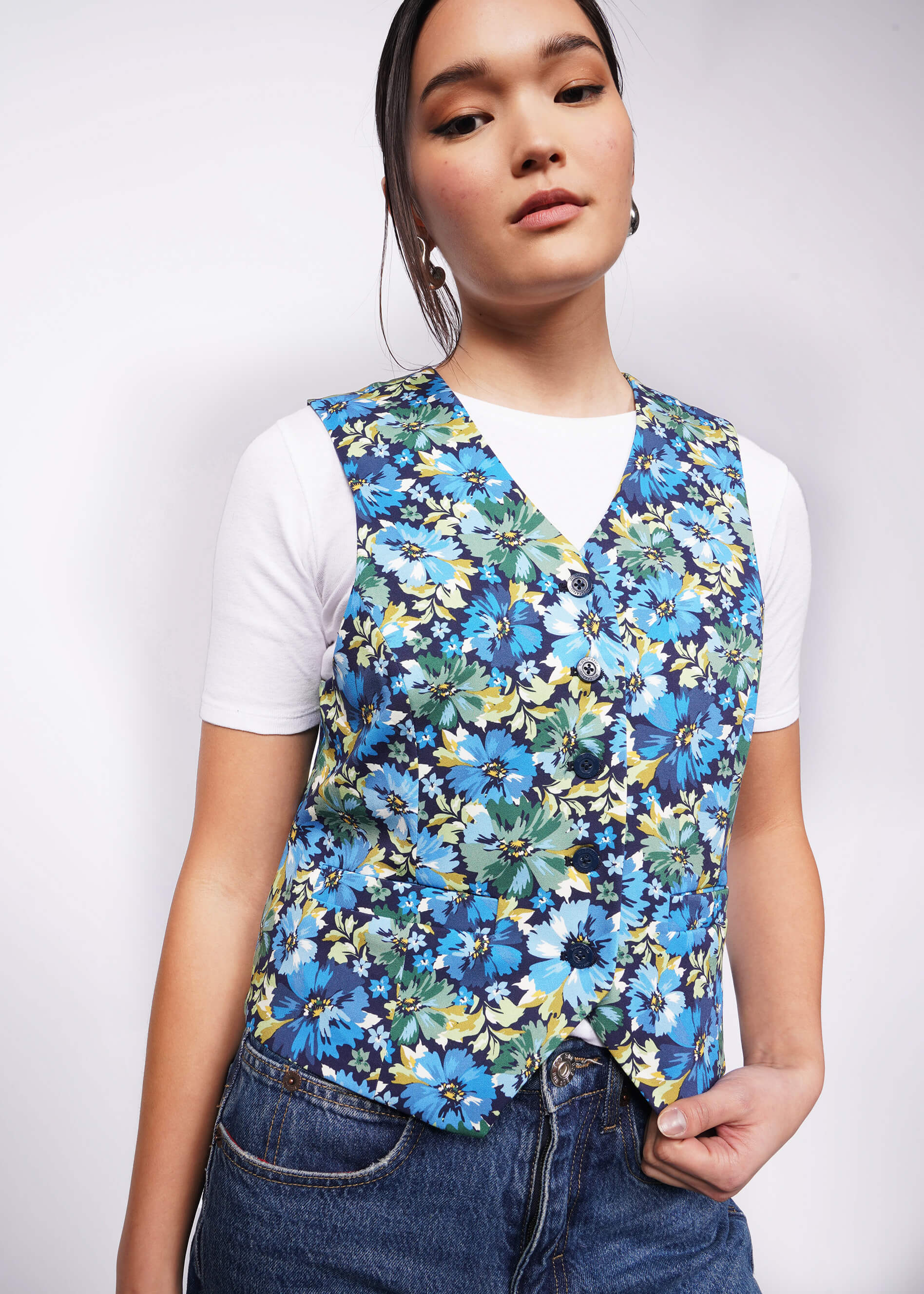 Person with black hair tied back looks at camera wearing white tshirt, jeans, and a floral patterned vest.