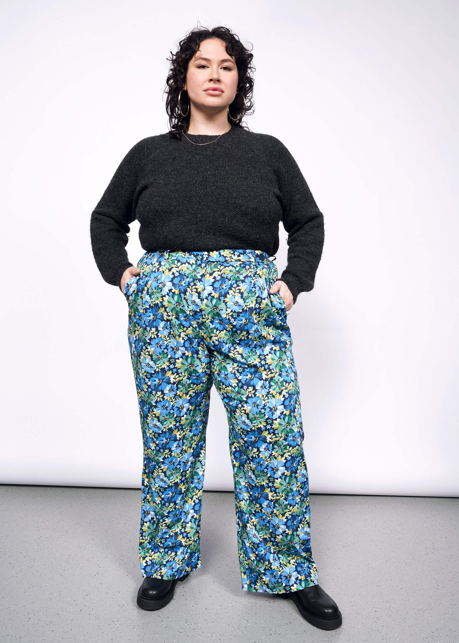 Person with short black curly hair wears floral pants and a black sweater and stands confidently looking at camera, showcasing her outfit against a neutral background.