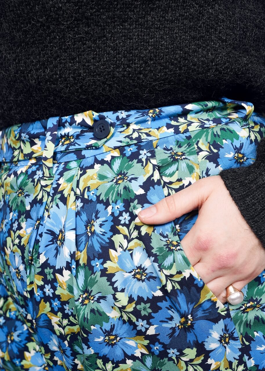 Close up of waistband detail of blue, green, and yellow floral patterned wide leg trouser. Model's hand is in pocket.