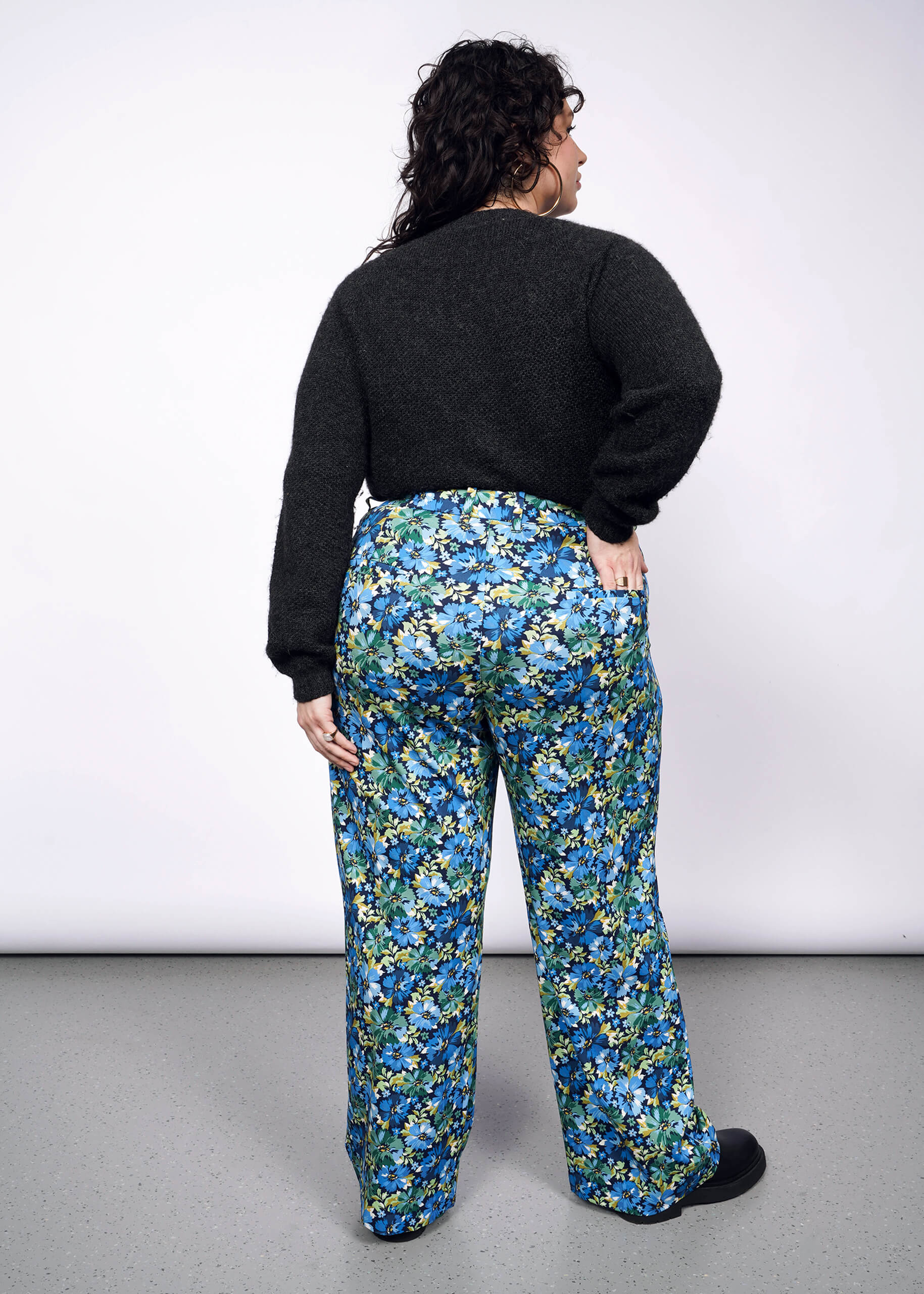 Back view of person with curly short black hair wearing black sweater tucked into patterned floral wide leg trousers and black boots.