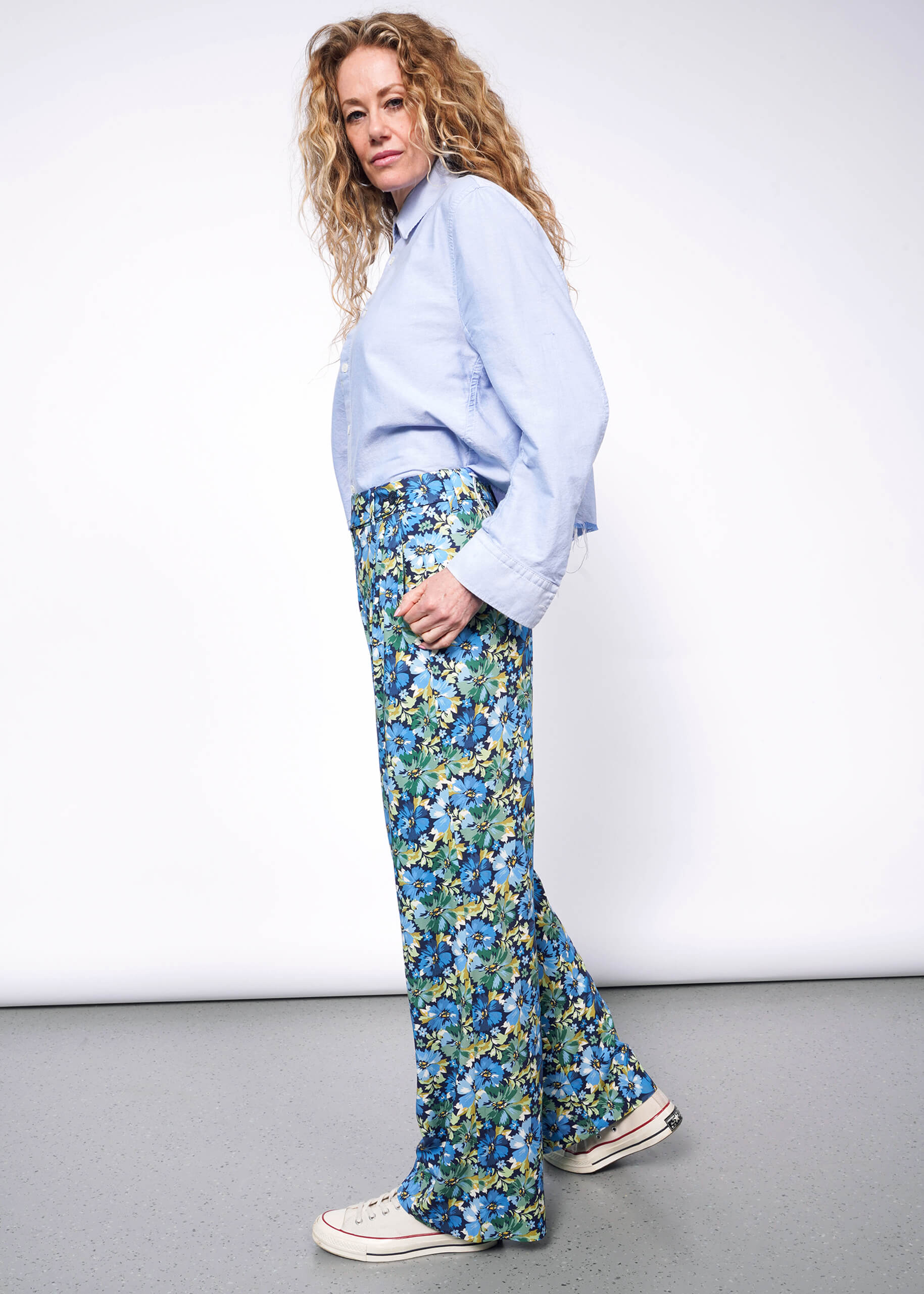 Side view of person with medium length wavy blonde hair looking at camera wearing light blue button up shirt tucked into patterned floral blue and green wide leg trousers and Converse sneakers. Neutral background.