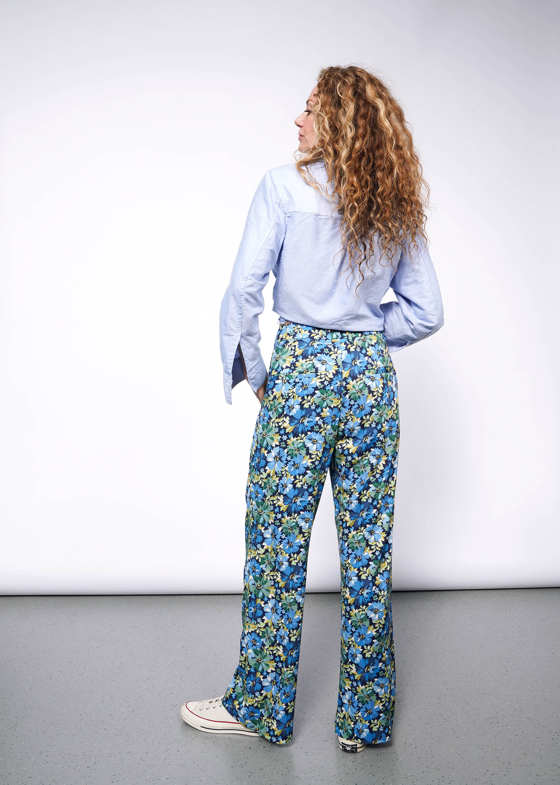 Back view of person with medium length wavy blonde hair looking to the left wearing light blue button up shirt tucked into patterned floral blue and green wide leg trousers and Converse sneakers. Neutral background.