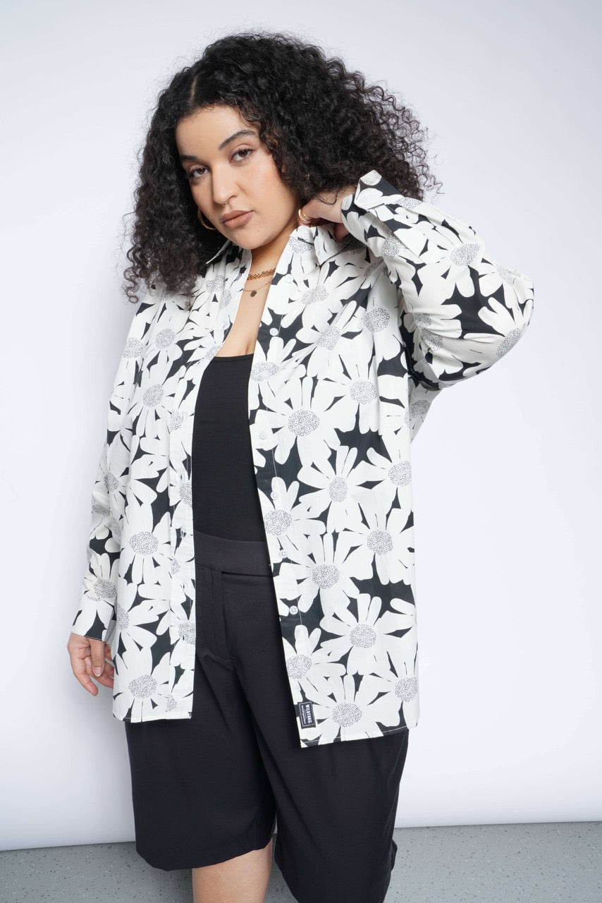 A person with curly hair confidently poses in a black top featuring The Essential Convertible Sleeve Button Up—loose-fitting, floral-patterned cotton—and pairs it with black shorts, against a plain background.