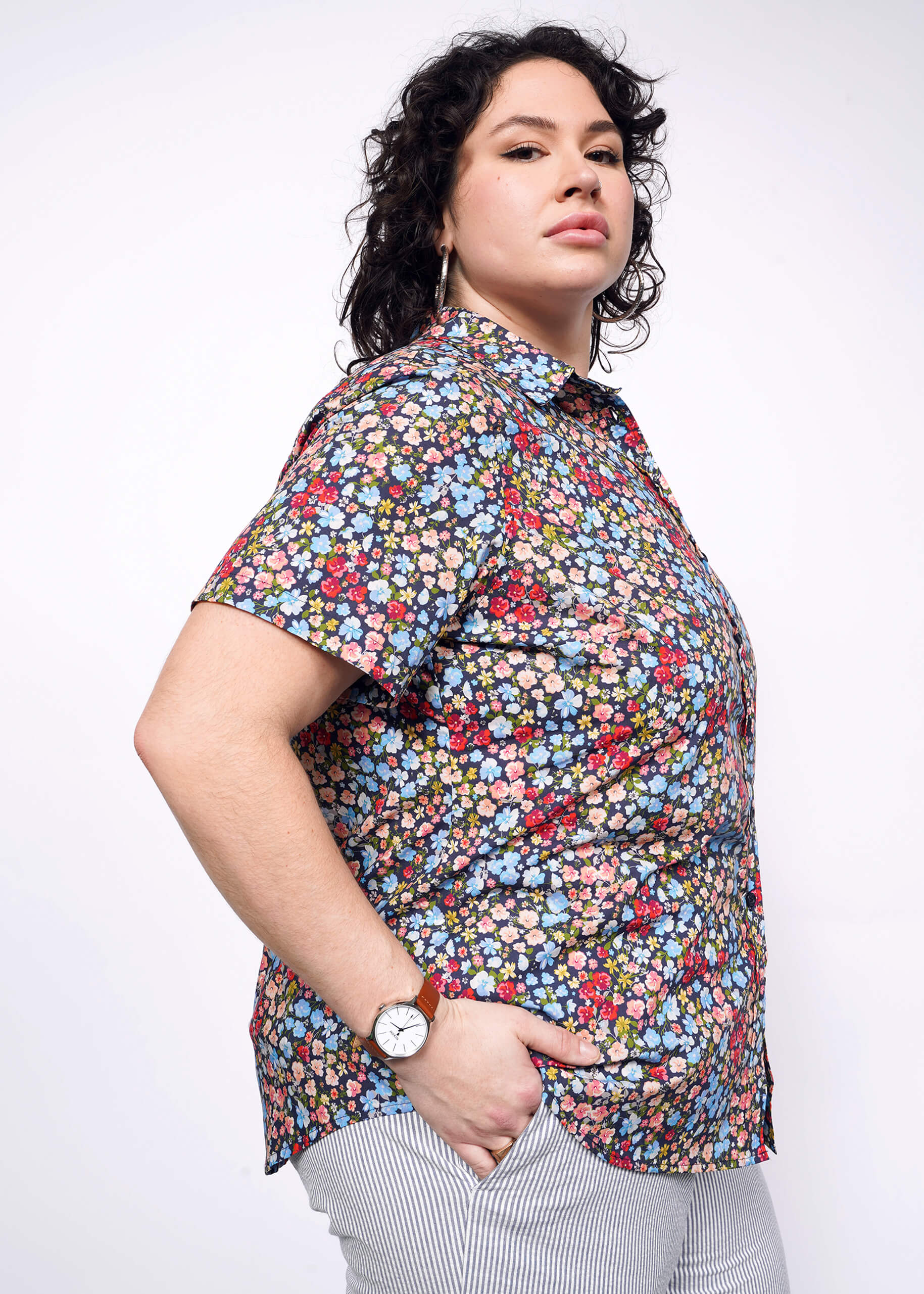 Side view of person with short black curly hair with hand in pocket wearing seersucker pants and flower printed short sleeve button up shirt.