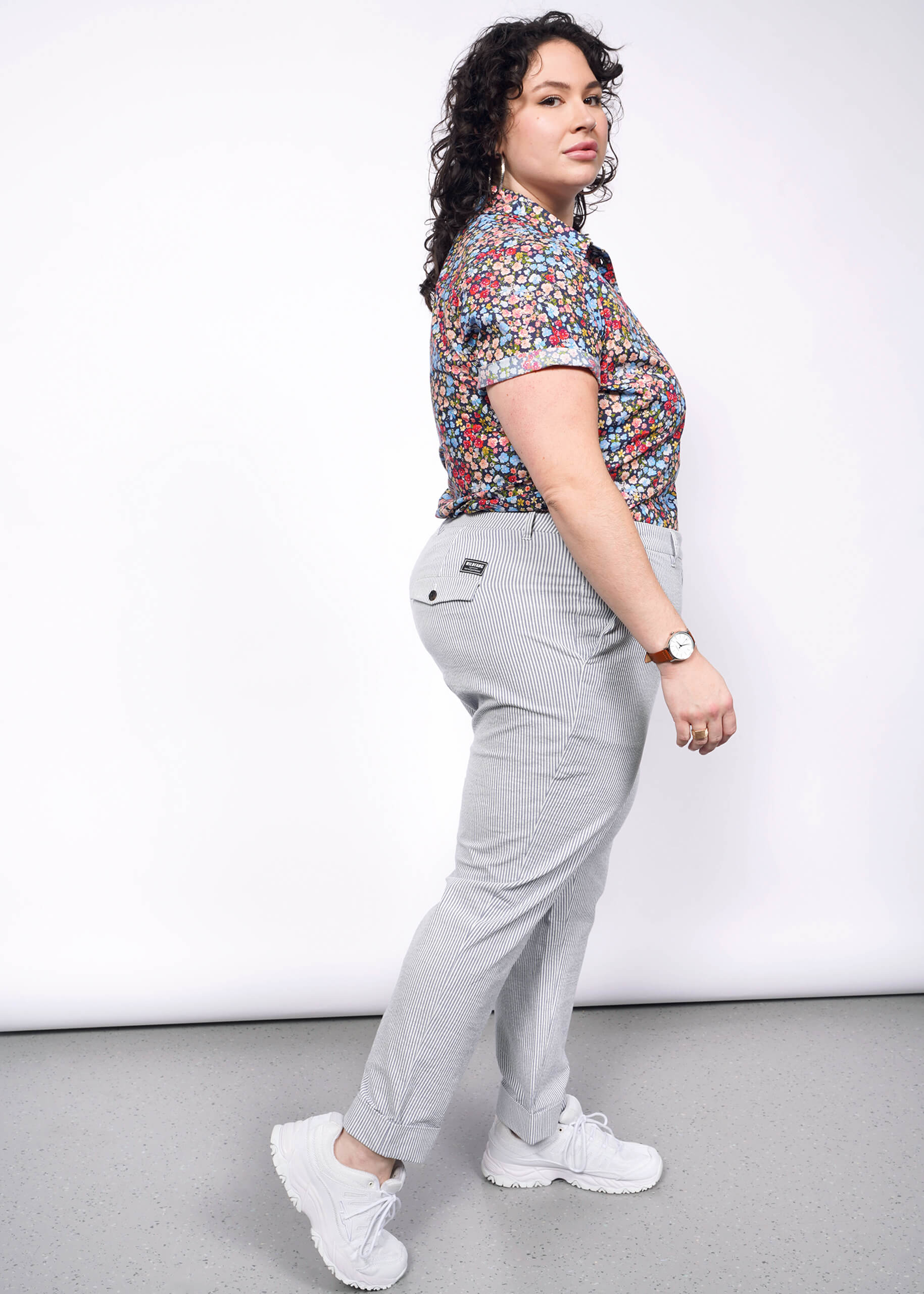 Side view of person with short black curly hair wearing seersucker pants, white sneakers, and flower printed short sleeve button up shirt.