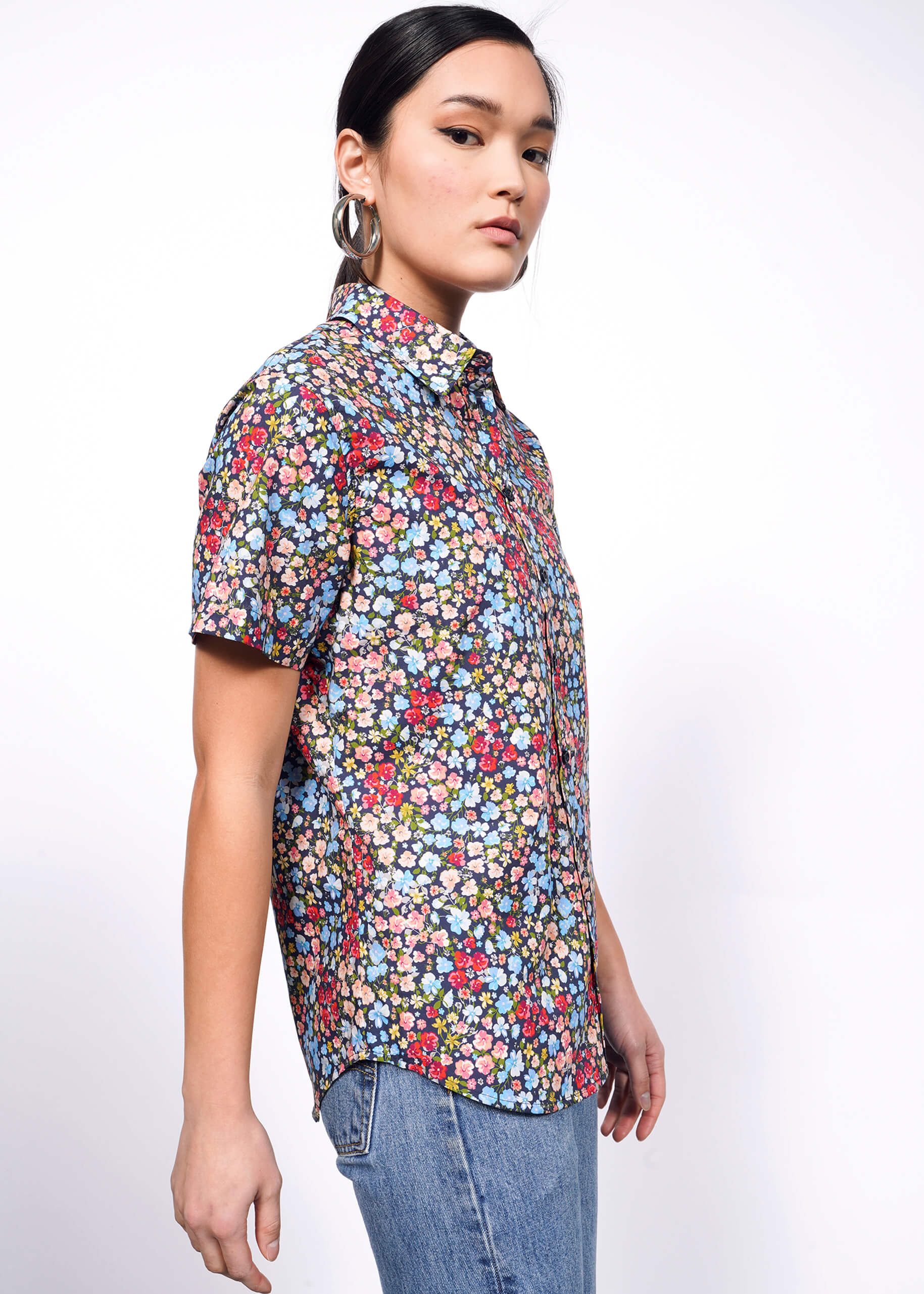 Side view of person with black hair tied back looking at camera wearing hoop earrings, jeans, and flower printed short sleeve button up shirt.