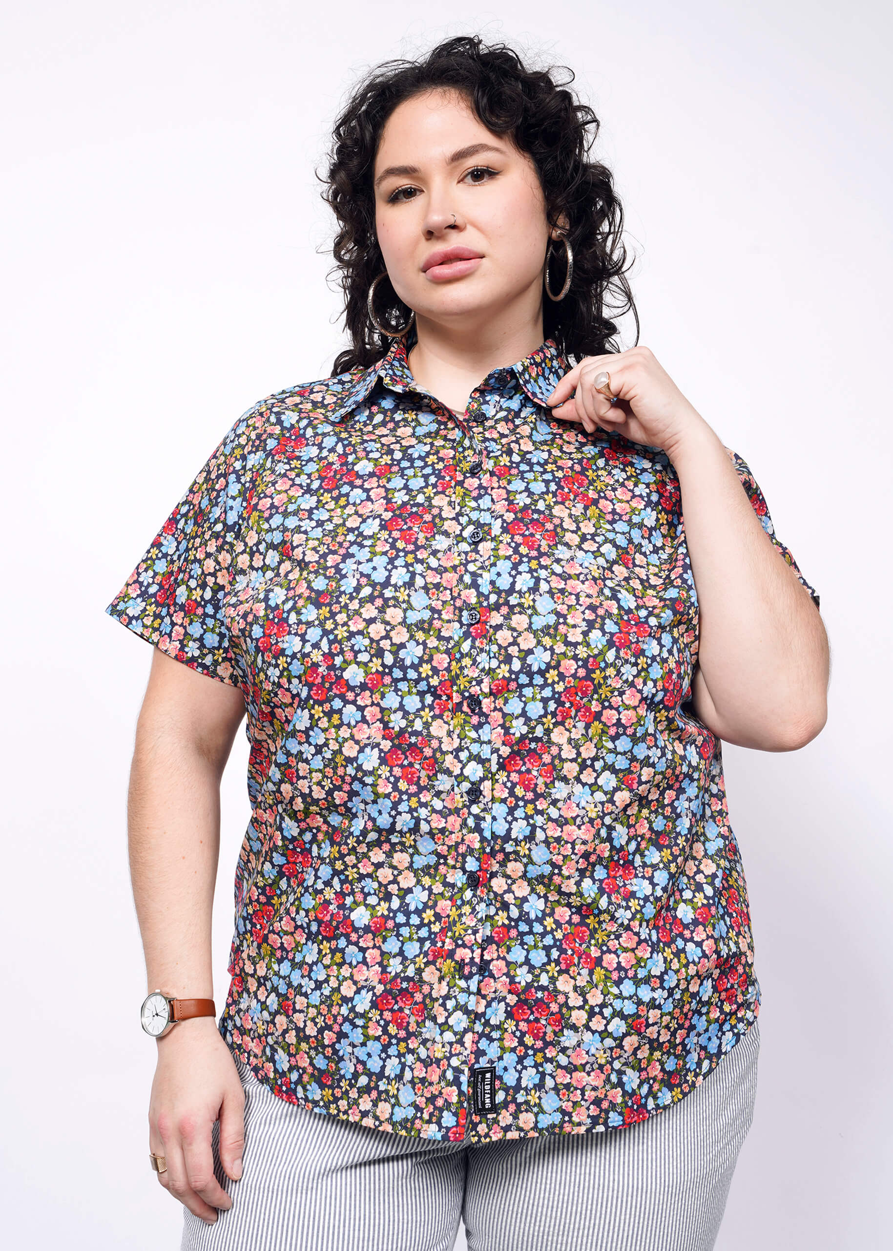 Person with short black curly hair holds collar of flower printed short sleeve button up shirt.