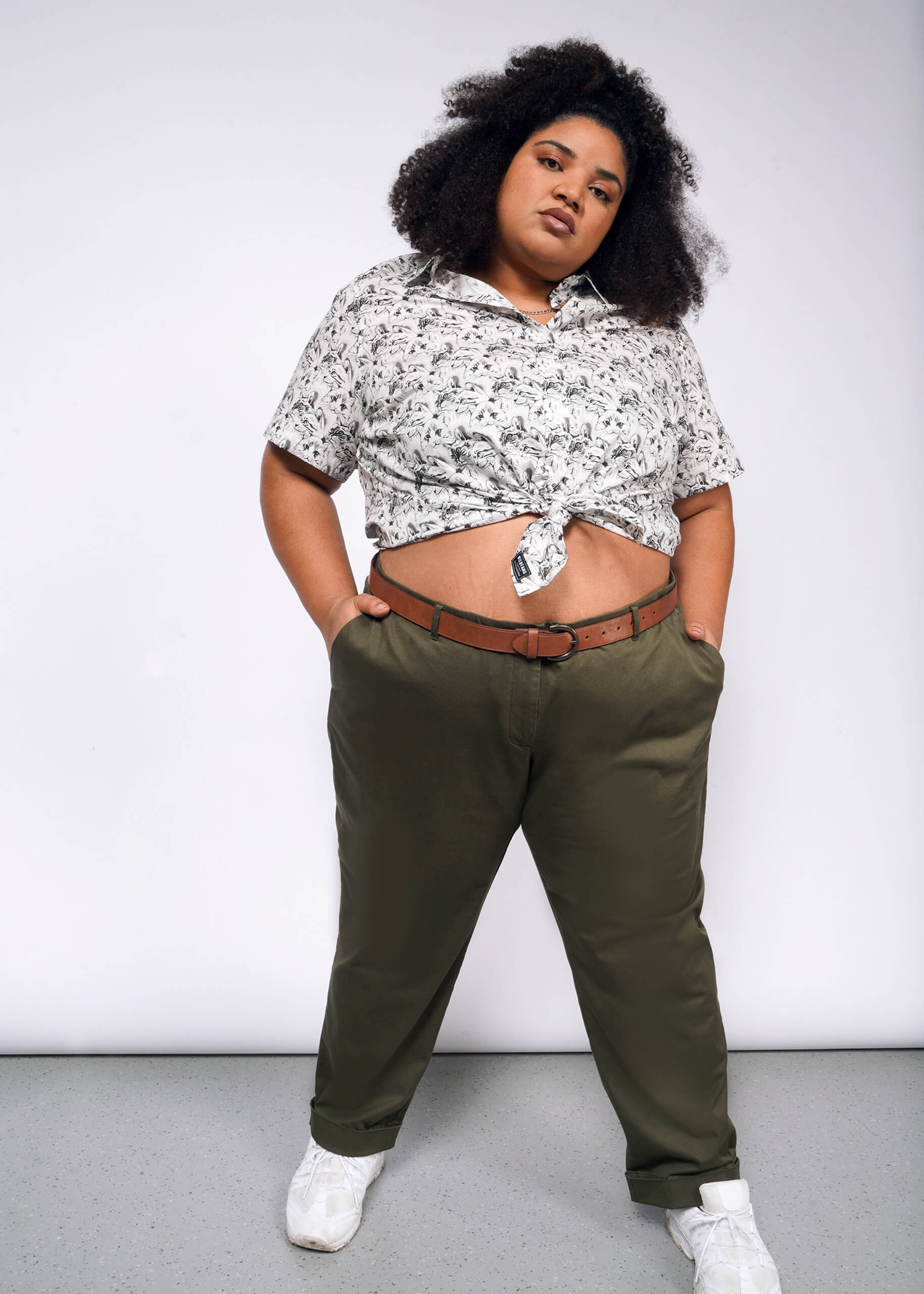 A person with curly hair stands confidently, hands in pockets, wearing The Essential Button Up—a bold print, short-sleeve shirt with Wildfang branded buttons and tied at the waist. Its paired with olive green pants and white sneakers, standing out against a plain gray background.