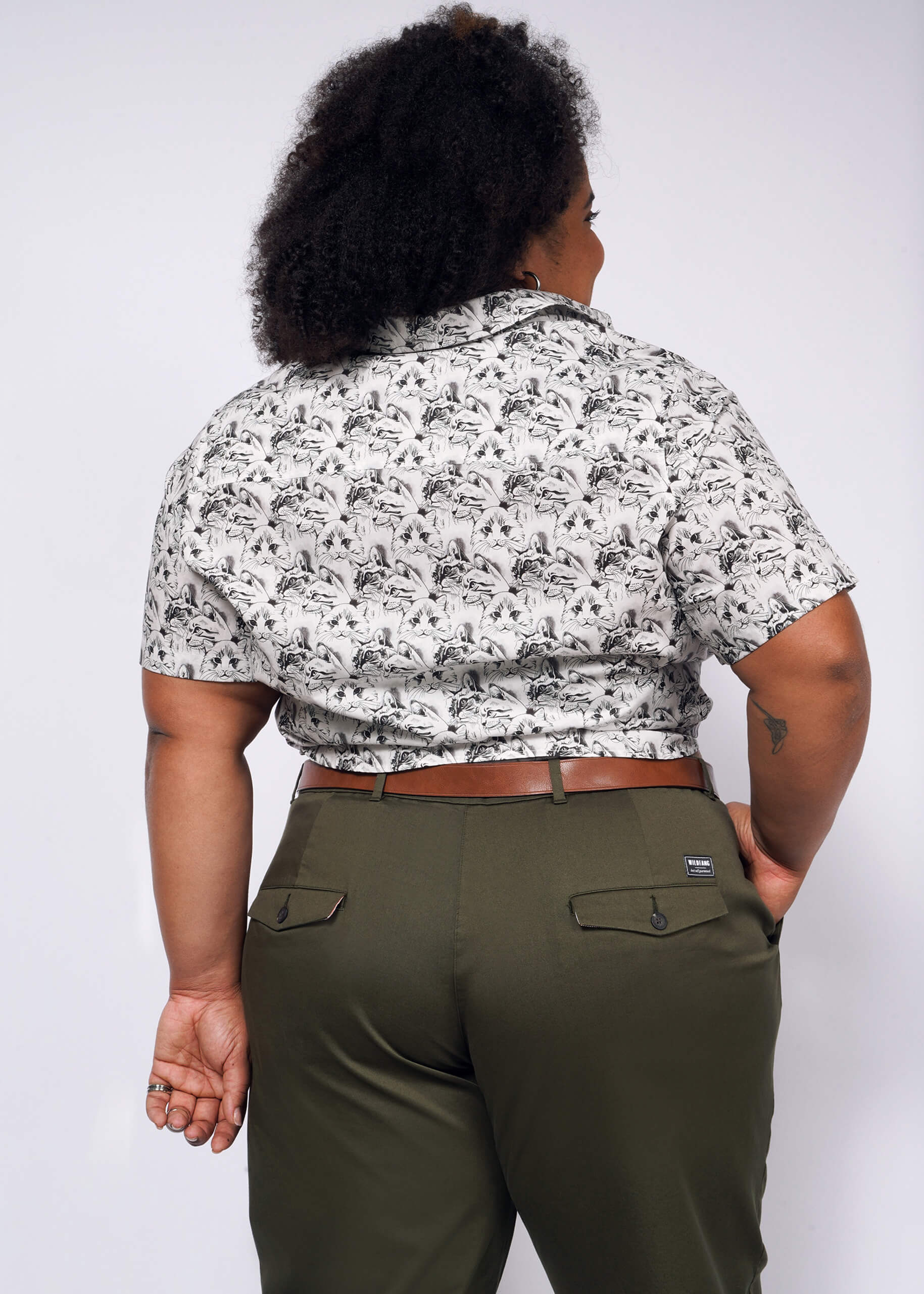 Facing away, the curly-haired individual wears The Essential Button Up with bold prints and olive green pants. Their left hand rests by their side against a plain backdrop, featuring Wildfang branded buttons for a touch of tailored fit elegance.