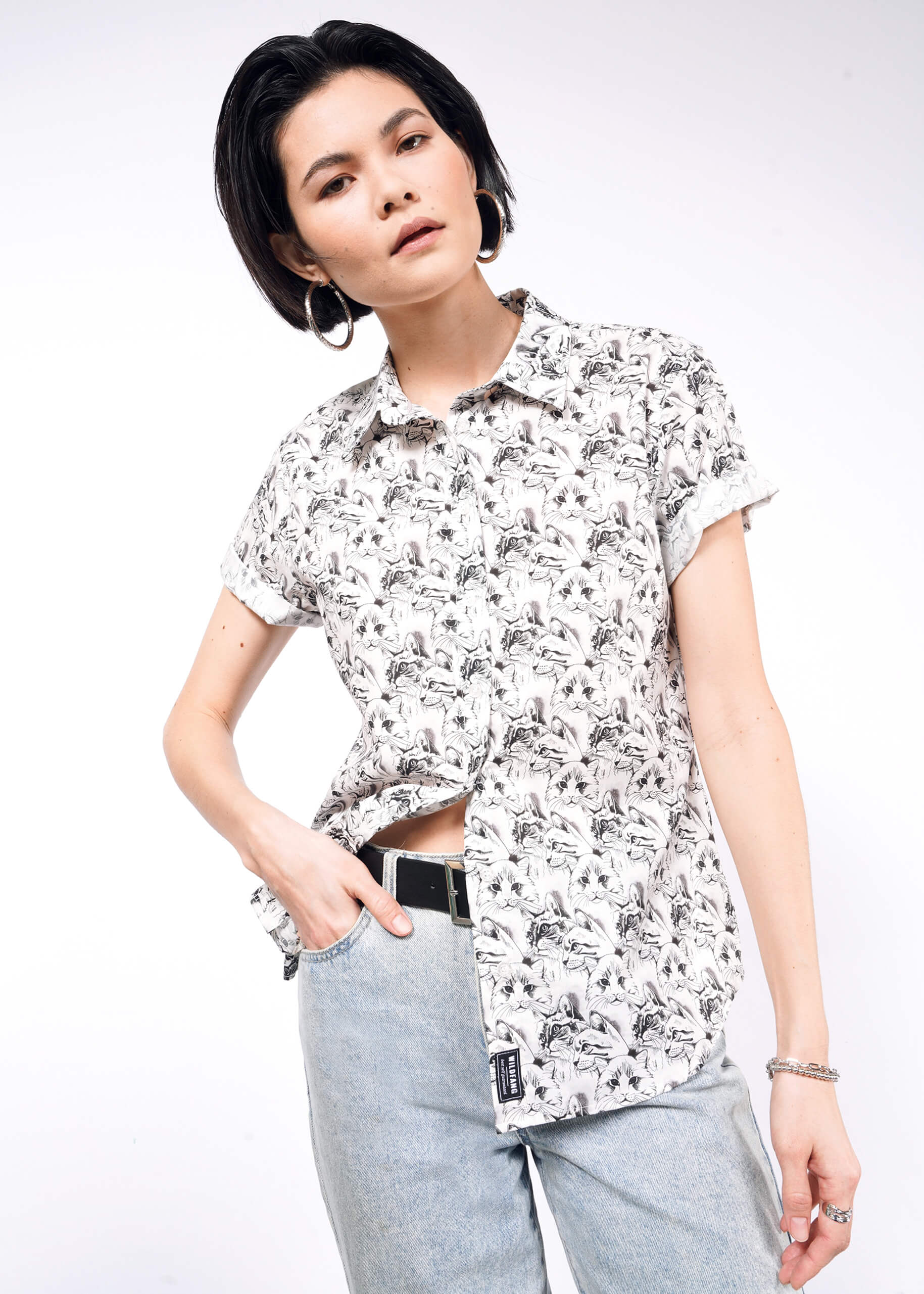 A person with short dark hair poses against a white background, wearing The Essential Button Up, featuring bold prints and light jeans, with one hand in their pocket.