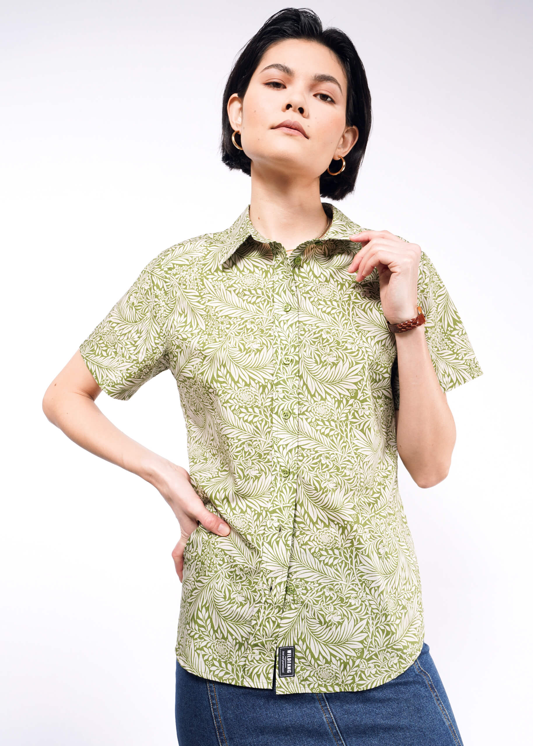 A person confidently poses against a white backdrop, wearing The Essential Button Up, featuring a green leafy pattern on breathable cotton, paired with blue denim jeans. With short dark hair and accessorized by hoop earrings and a bracelet, they embody classic Wildfang style.