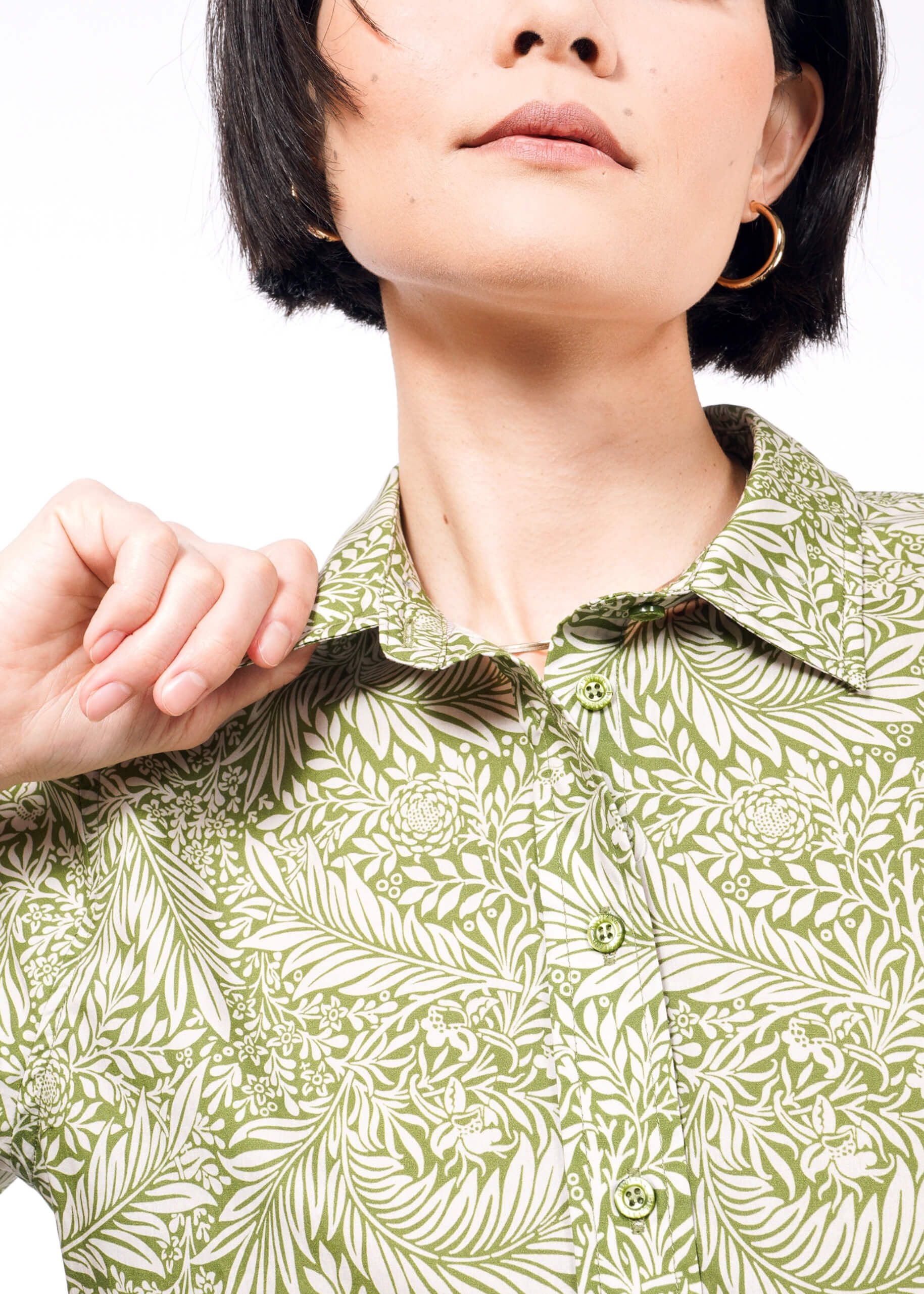 A woman with short black hair and hoop earrings wears a green shirt with white floral patterns, made from breathable cotton. Holding the tailored collar, she epitomizes The Essential Button Up by Wildfang against a plain white backdrop.