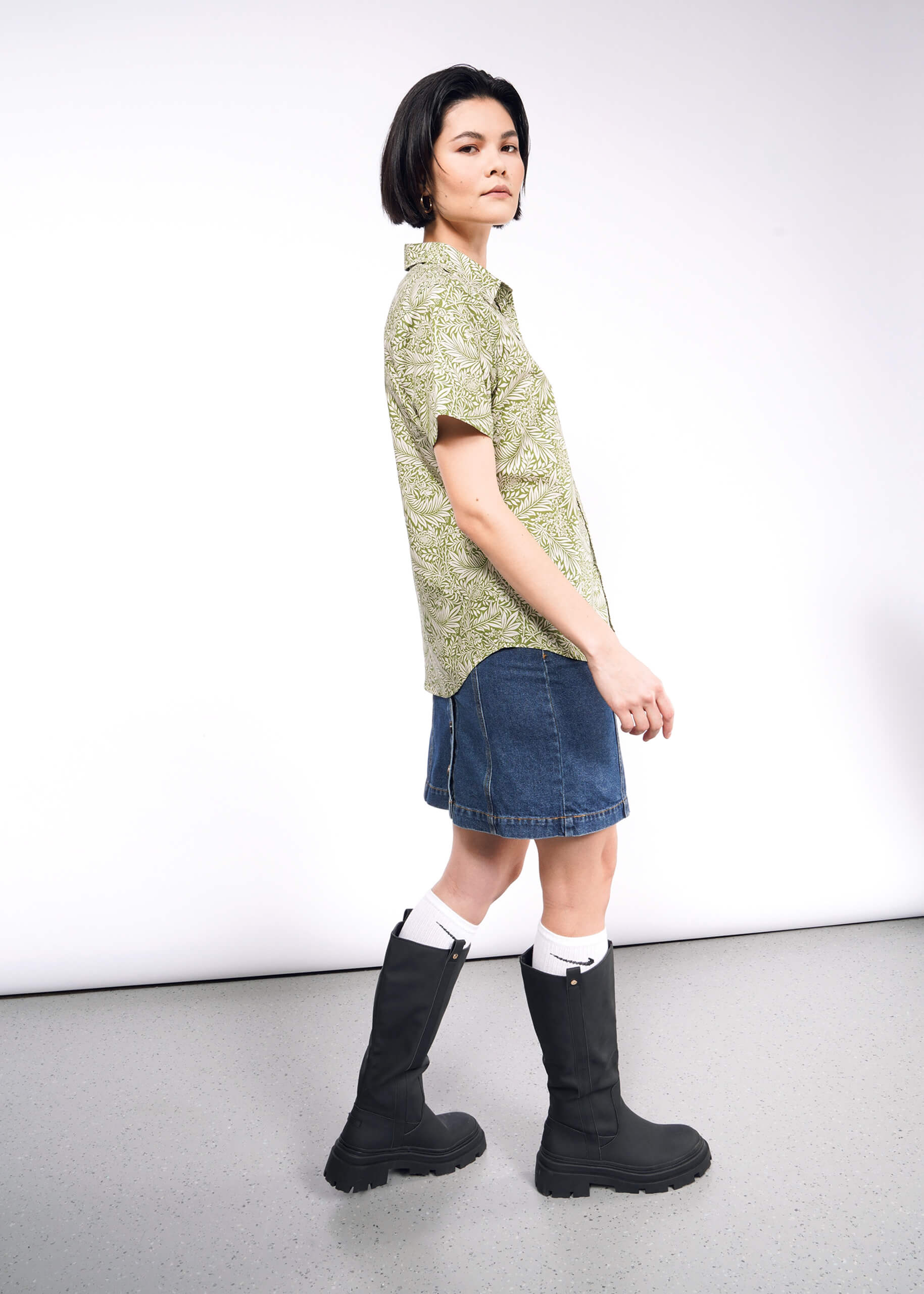 A person with short hair sports The Essential Button Up in a green patterned, tailored fit. They pair it with a denim skirt, knee-high black boots, and white socks while standing sideways against a plain background, facing the camera.