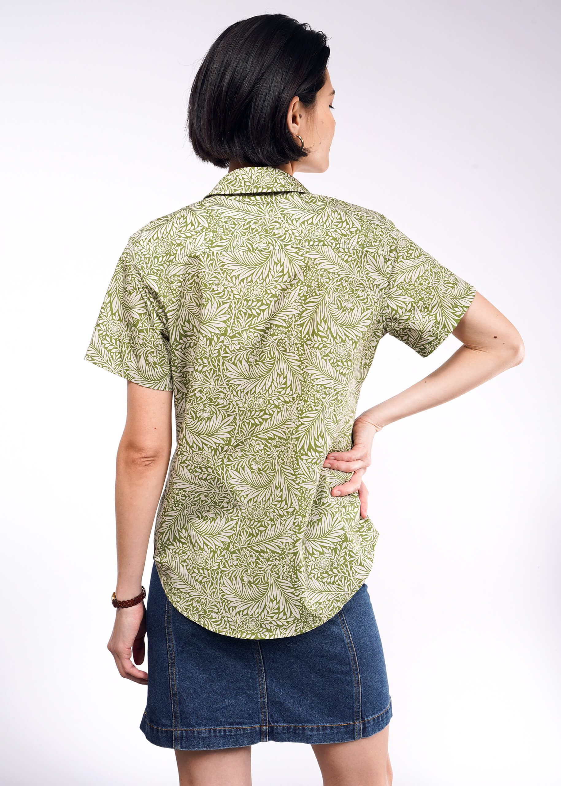 A person with short dark hair stands with their back to the camera, hand on hip, wearing a light green patterned short-sleeve The Essential Button Up in breathable cotton and a denim skirt against a plain white background.