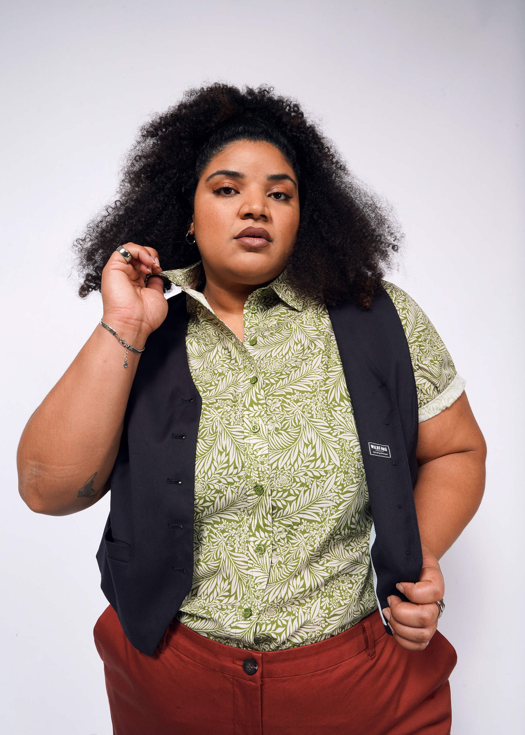 A person with curly hair showcases a Wildfang classic look, wearing The Essential Button Up in a green pattern, made of breathable cotton. They pair it with a tailored-fit black vest and rust-colored pants, confidently holding the shirts collar against a plain white background.