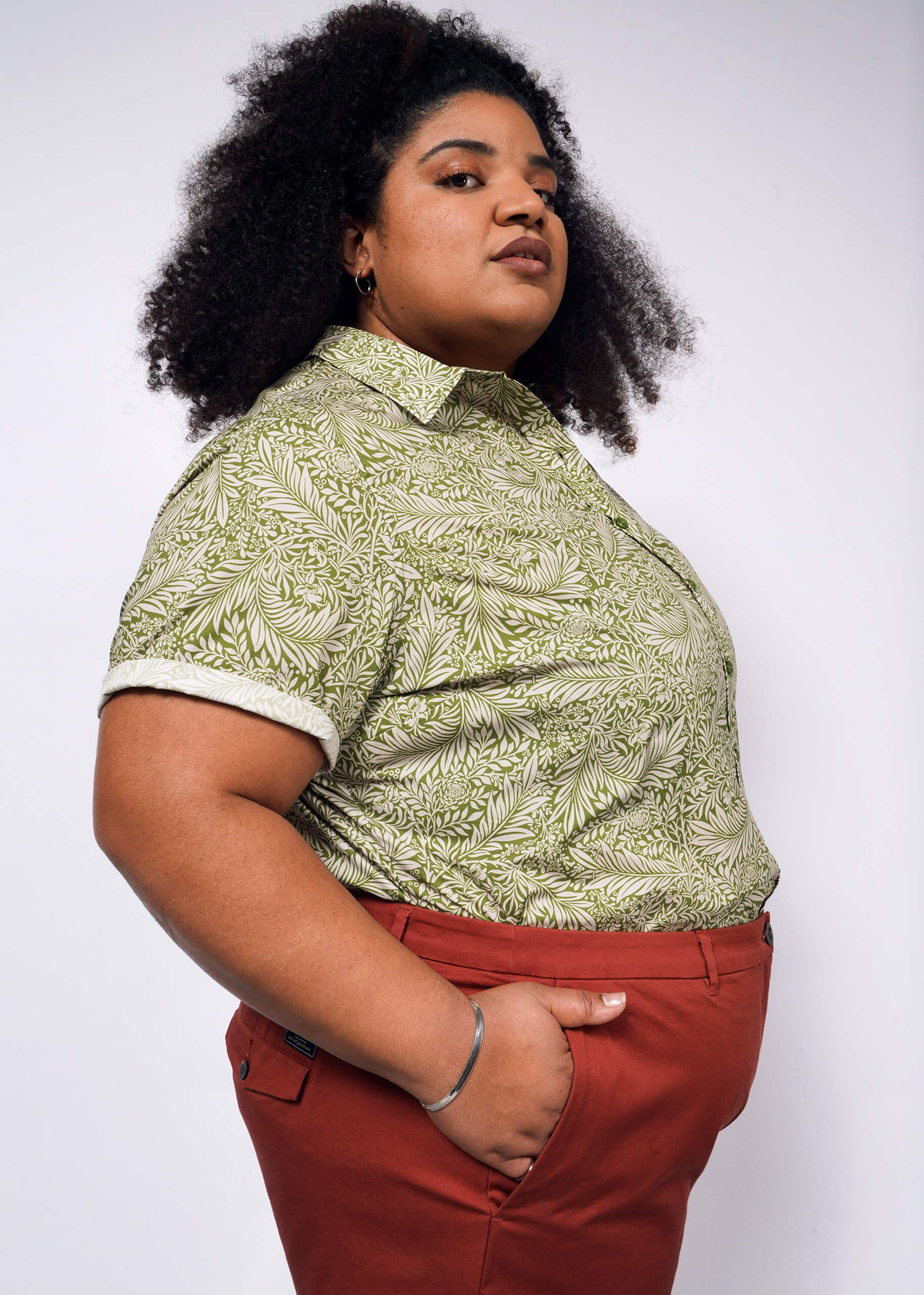 A person with curly hair confidently wears a green Wildfang The Essential Button Up and red pants. Their tailored fit enhances a serious expression against a plain background, exuding style and strength in breathable cotton.