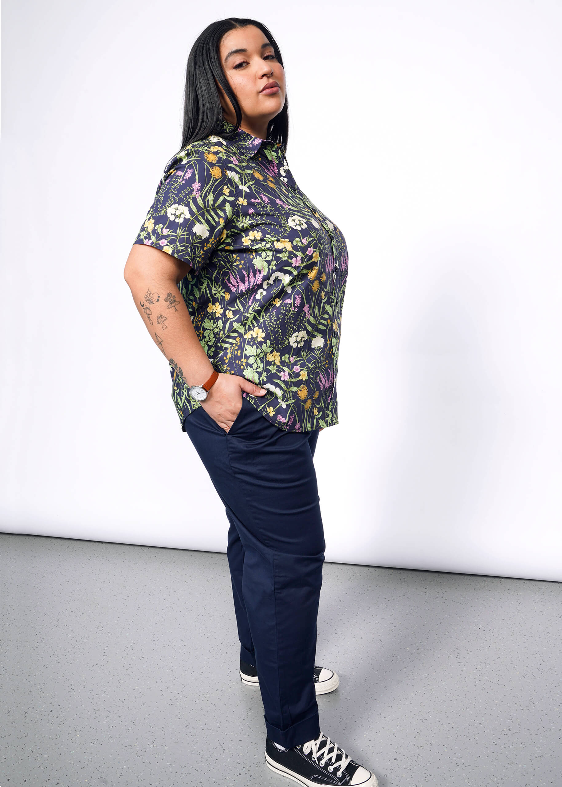 Side view of person with long black hair looking at camera with hand in pant pocket wearing navy trouser, Converse sneakers, and short sleeve button up shirt with botanical print.