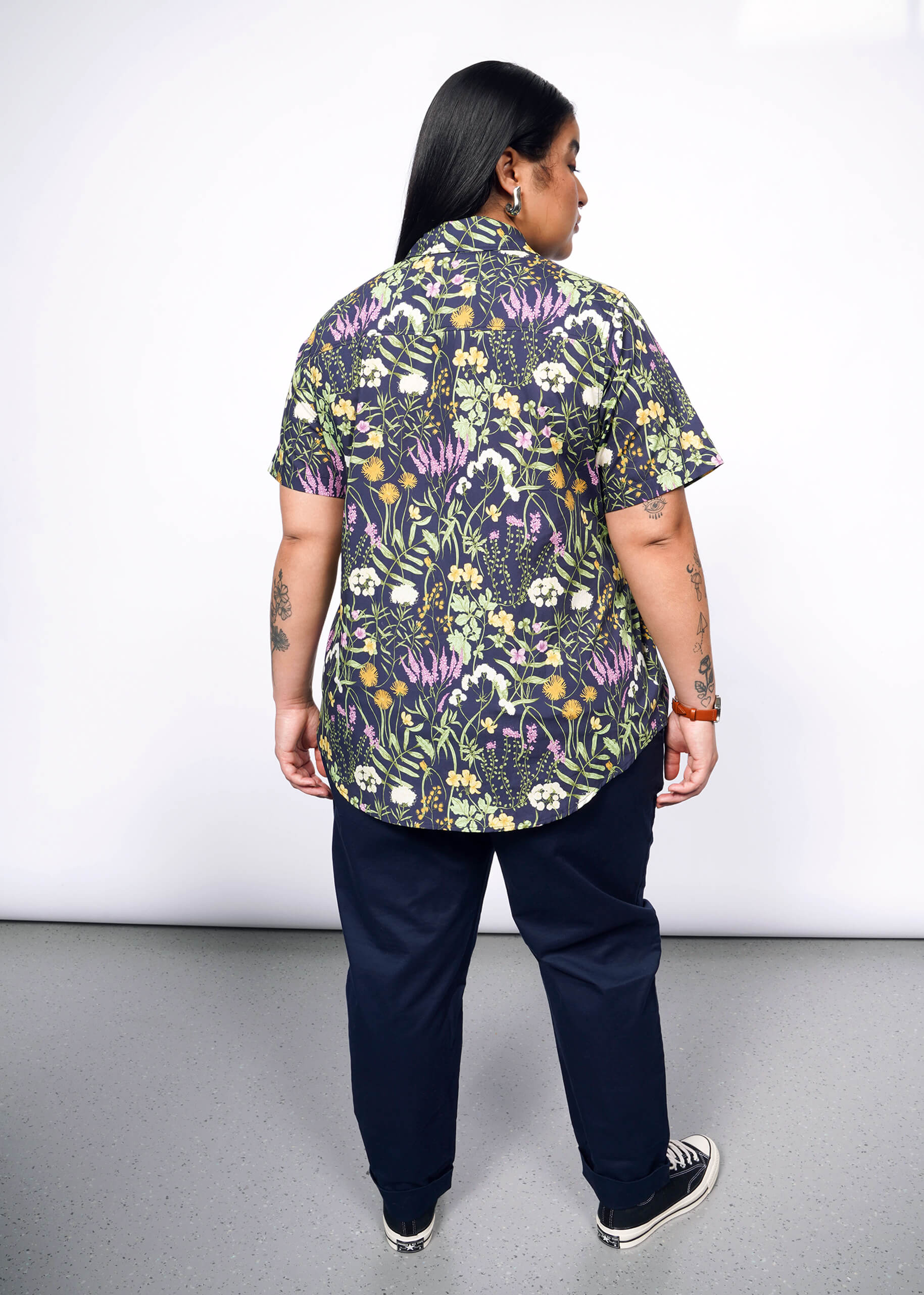 Back view of person with long black hair looking at camera wearing navy trouser, Converse sneakers, and short sleeve button up shirt with botanical print.