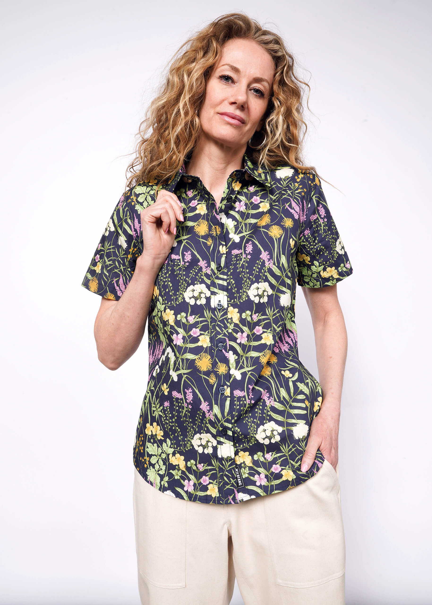 Person with wavy blonde hair holds collar of short sleeve botanical printed button up shirt.