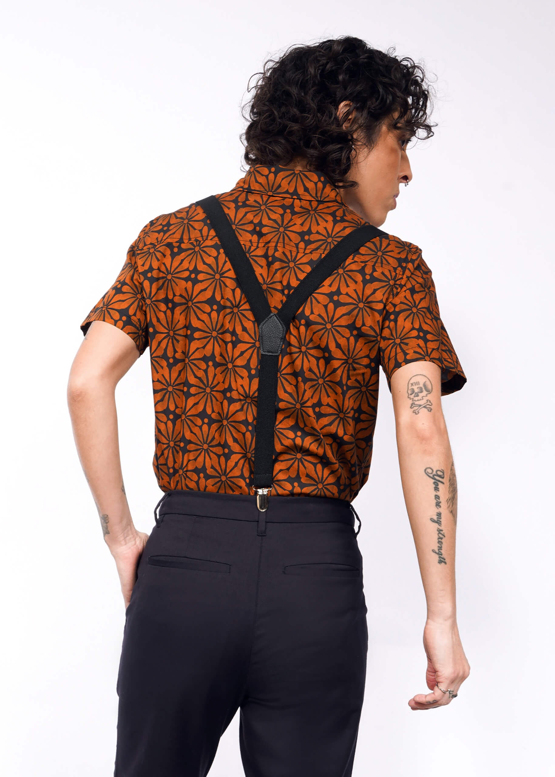 A person with curly hair wears The Essential Button Up, an orange short-sleeved shirt with a black geometric pattern. It drapes over their tattooed arms, paired with suspenders and dark pants against a plain white background, offering breathable comfort in cotton fabric.