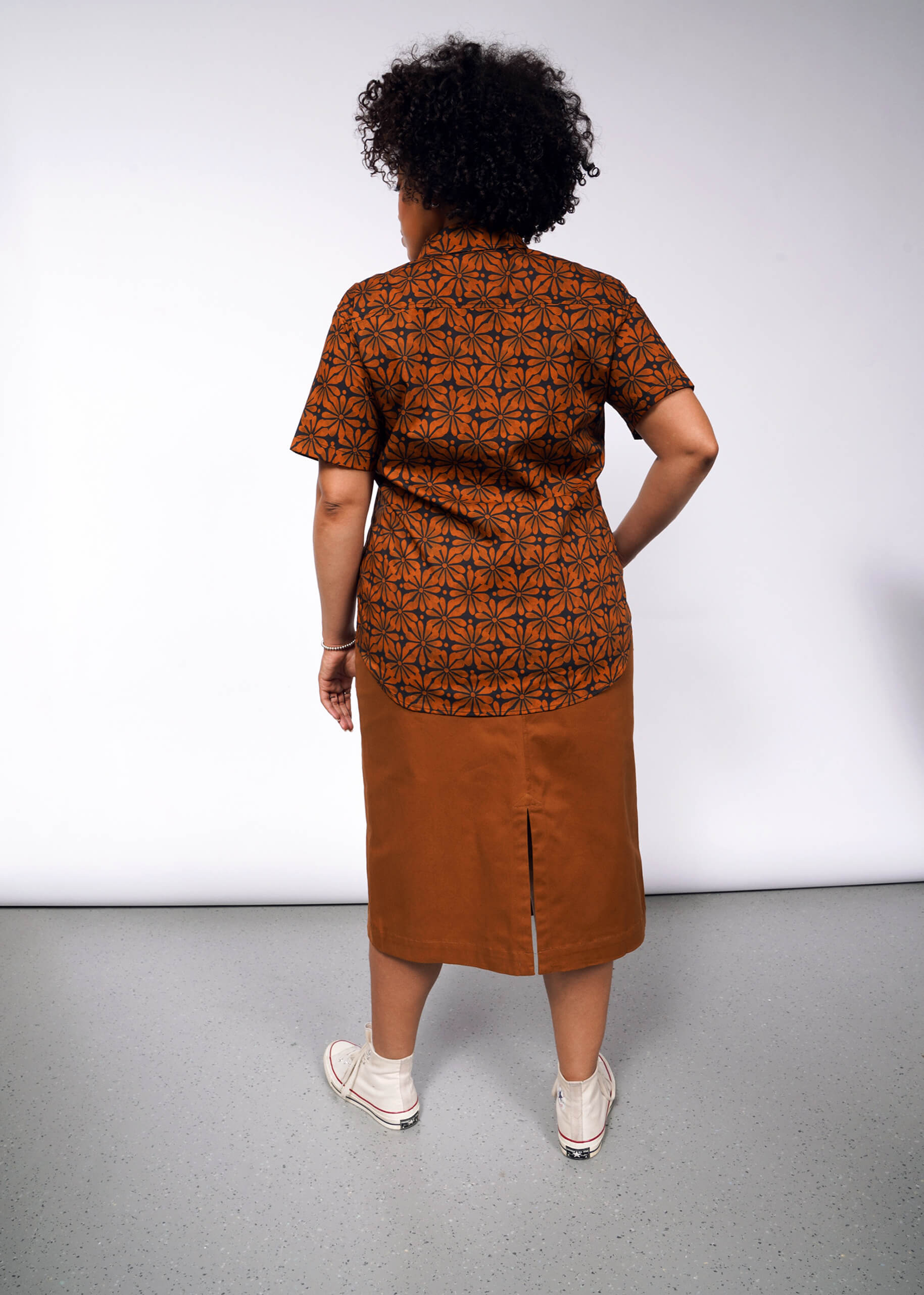 A person with curly hair, turned away from the camera, showcases The Essential Button Up, a tailored patterned brown shirt paired with a knee-length brown skirt featuring a back slit. White shoes complete the look against a plain light gray wall.